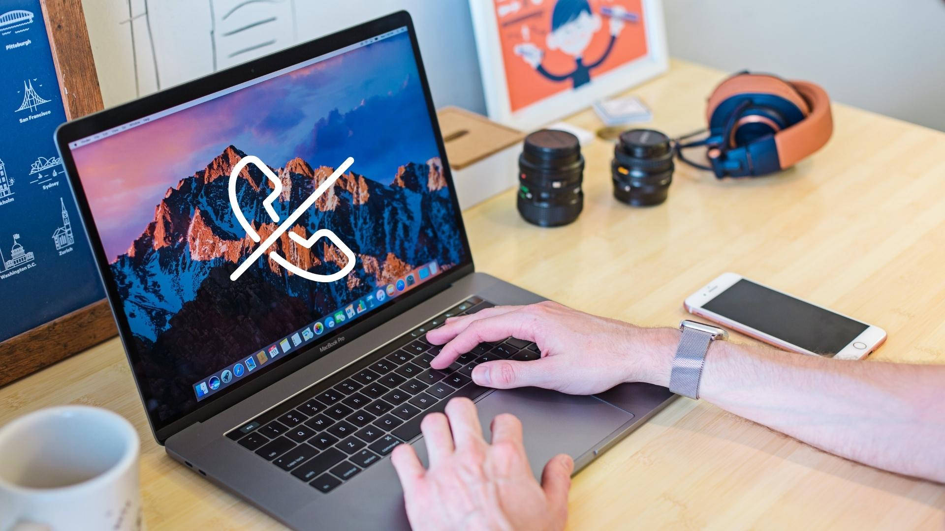 macbook 桌布 壁紙 — 一個人在一部顯示山脈桌布的手提電腦上打字，並在木桌上疊加了一個電話靜音圖標，桌上還有耳機和相機鏡頭。