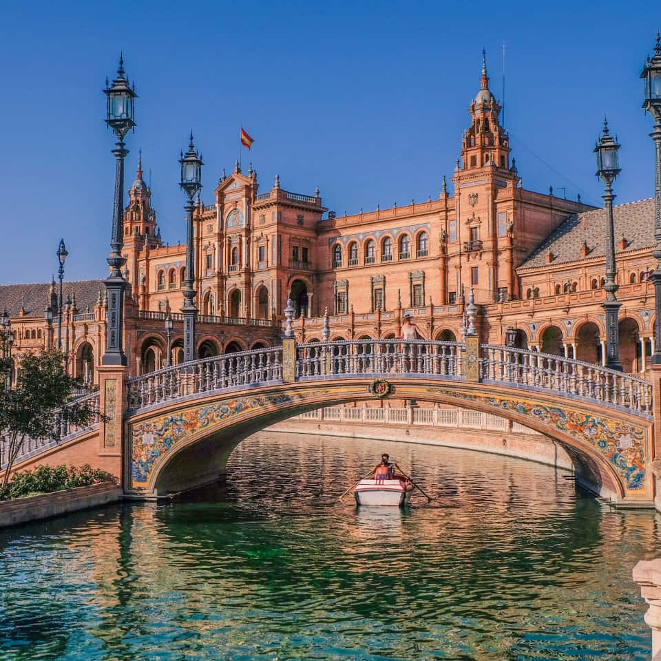 Barcelona 桌布 壁紙 — 一個人在塞維利亞宏偉的西班牙廣場前，在晴朗的藍天下，划著小船穿過色彩繽紛的瓷磚橋。