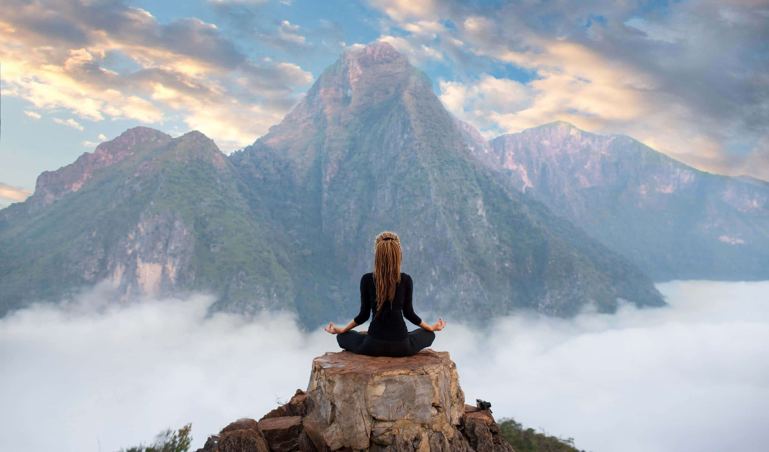 Concentration 桌布 壁紙 — 一個人在岩石山頂上冥想，下方雲霧繚繞，雄偉的山脈在柔和的金色天空下聳立。