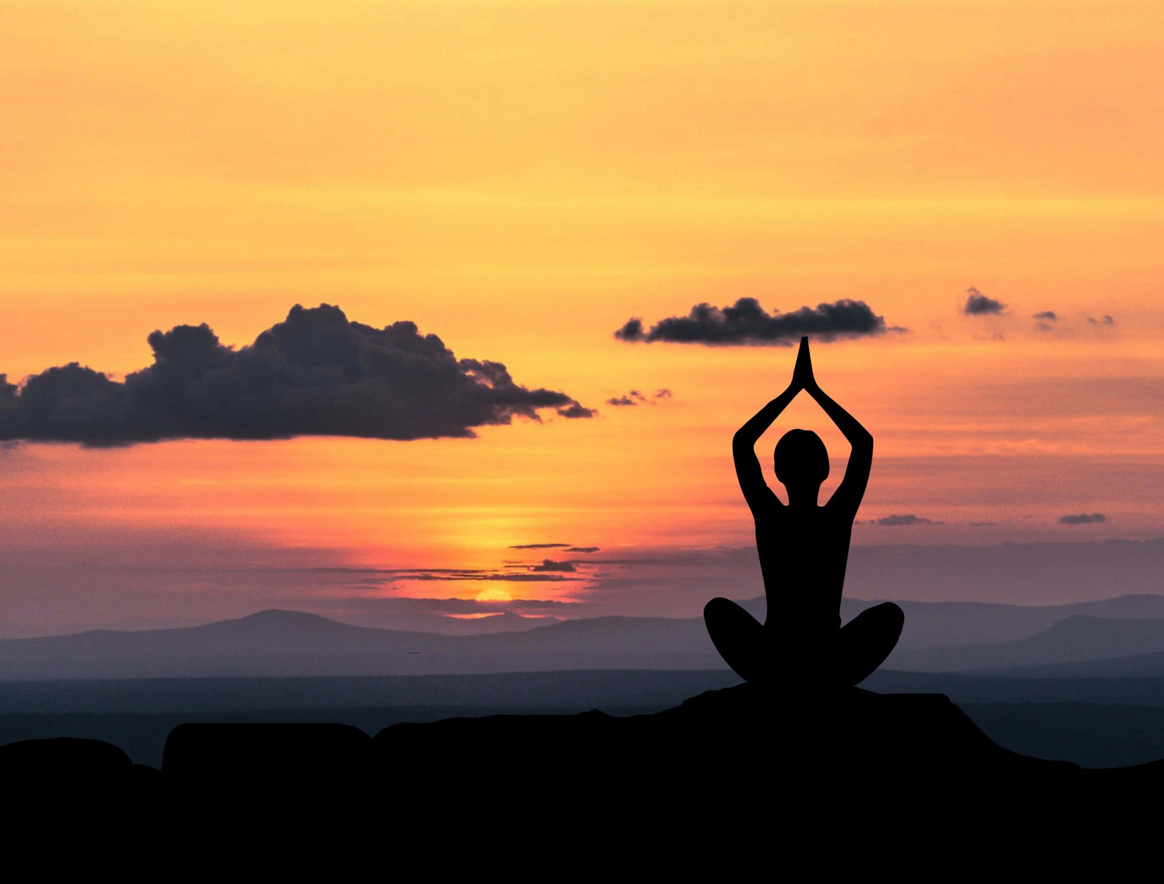Spiritual Meditation 桌布 壁紙 — 一個人在懸崖頂上做瑜伽姿勢的剪影，背景是夕陽下充滿活力的橙色和粉紅色天空以及遠處的山脈。