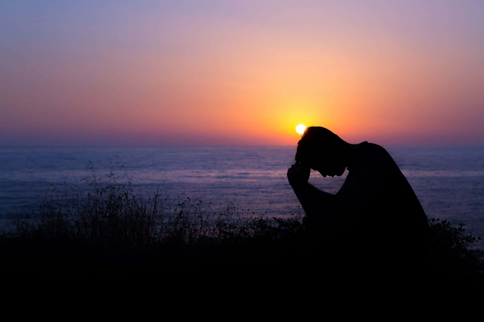 Praying 桌布 壁紙 — 一個人在日落時分沉思的剪影，俯瞰海洋，天空呈現鮮豔的橙色和紫色。