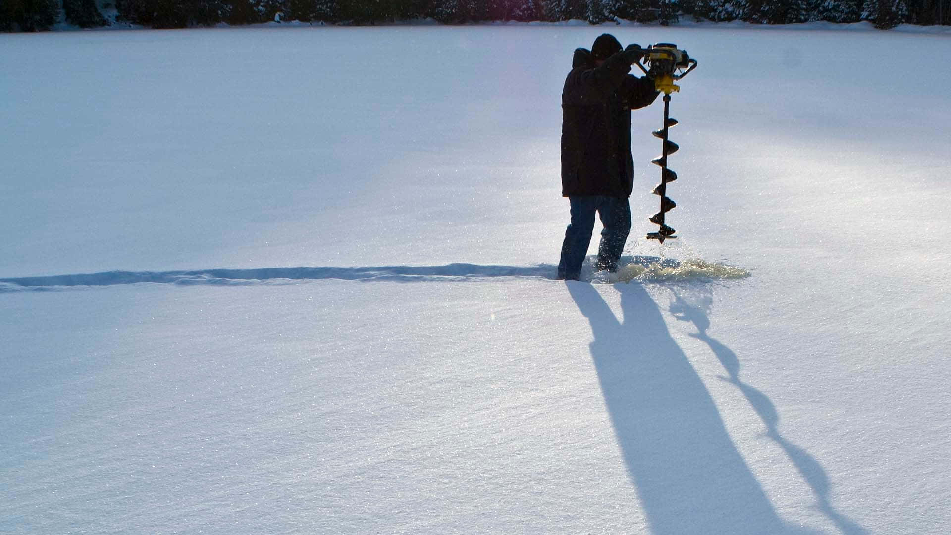 Ice Fishing 桌布 壁紙 — 一個人在明亮的冬日陽光下，在結冰的湖面上鑽穿厚冰，投下長長的影子。