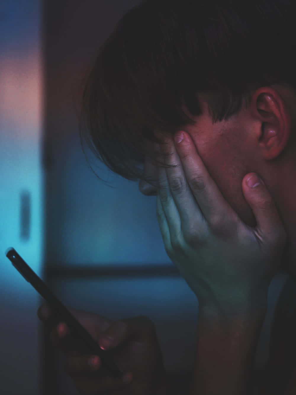憂鬱 桌布 壁紙 — 一個人在昏暗的藍光下用一隻手遮住臉，另一隻手拿著手機。