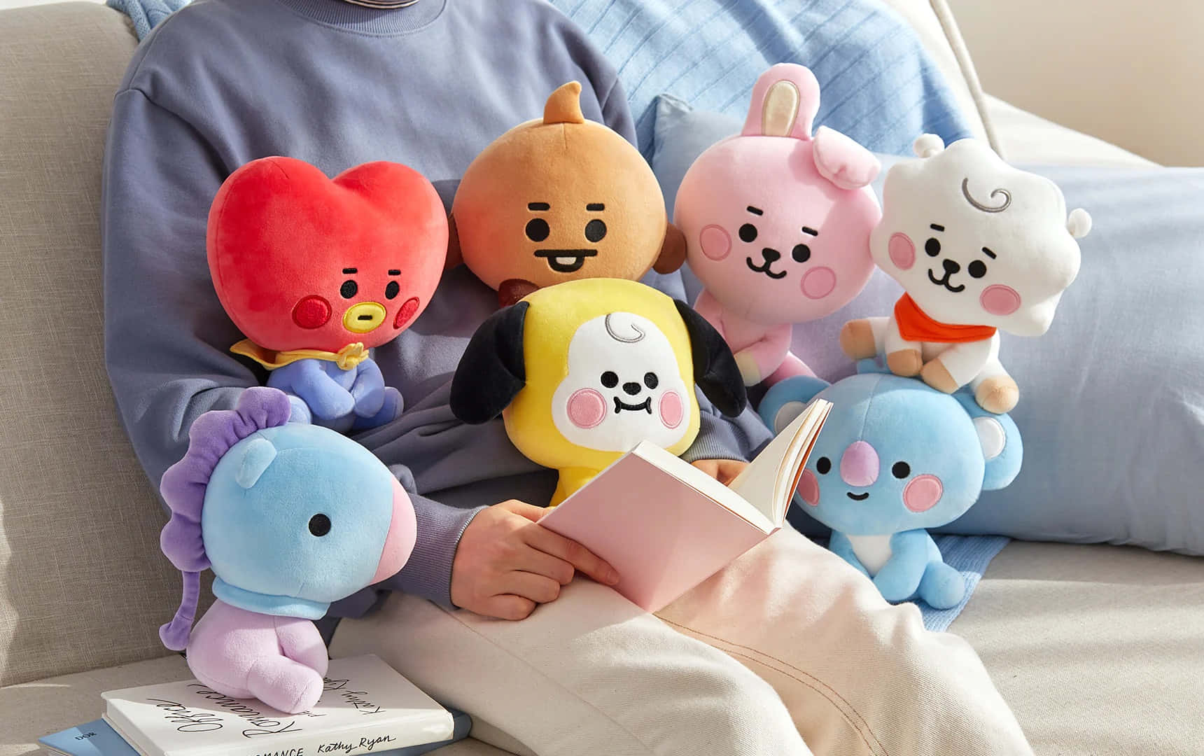 BT21 桌布 壁紙 — 一個人在沙發上放鬆，周圍擺滿色彩繽紛的BT21毛絨玩具，同時閱讀一本書。