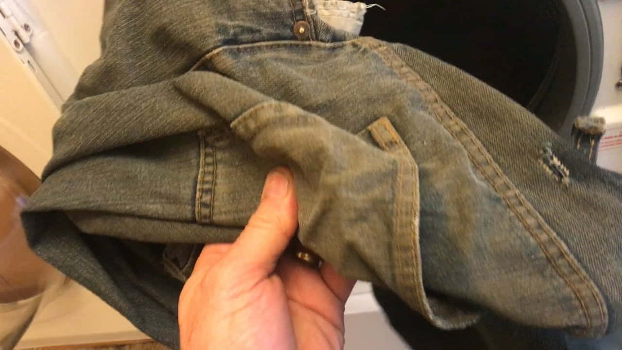 一個人在洗衣房拿著一雙牛仔褲 壁紙 — 一隻手拿著一條磨損的褪色藍色牛仔褲，邊緣磨損，鈕扣可見，背景是模糊的室內環境。