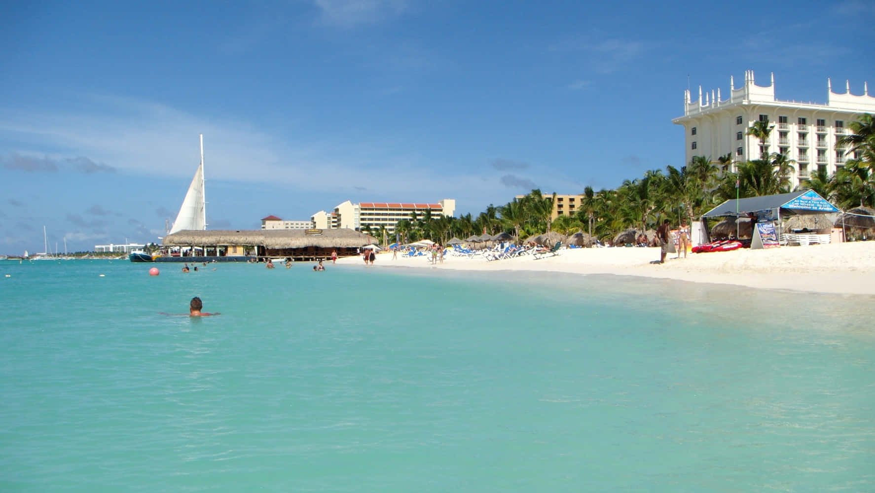 Aruba 桌布 壁紙 — 一個人在清澈碧綠的海水中游泳，靠近一條有棕櫚樹和度假村建築的白沙灘，天空湛藍明亮。