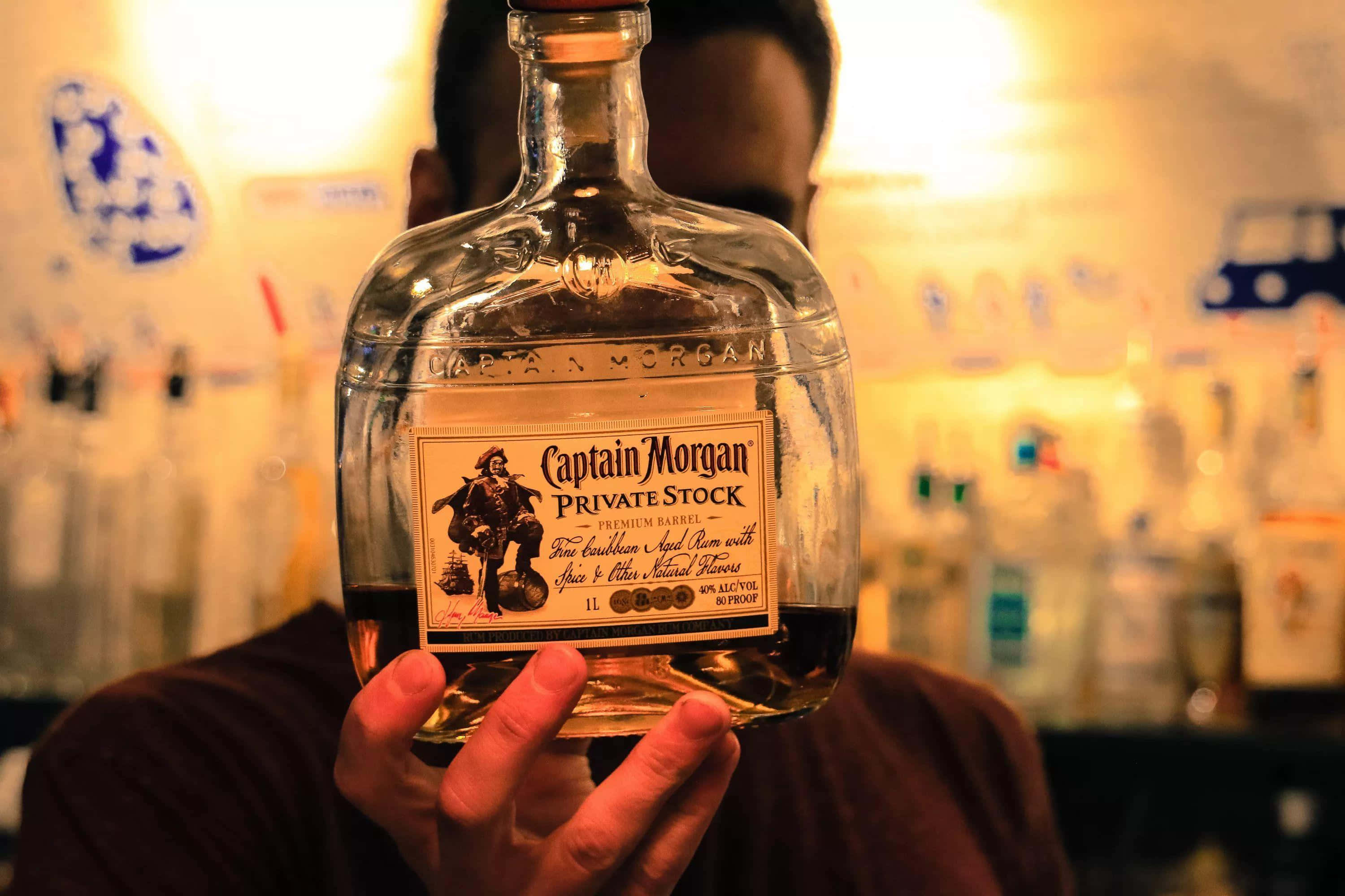 alcohol 桌布 壁紙 — 一個人在燈光溫暖的酒吧裡，手持一瓶Captain Morgan Private Stock冧酒，身後是模糊的酒架。