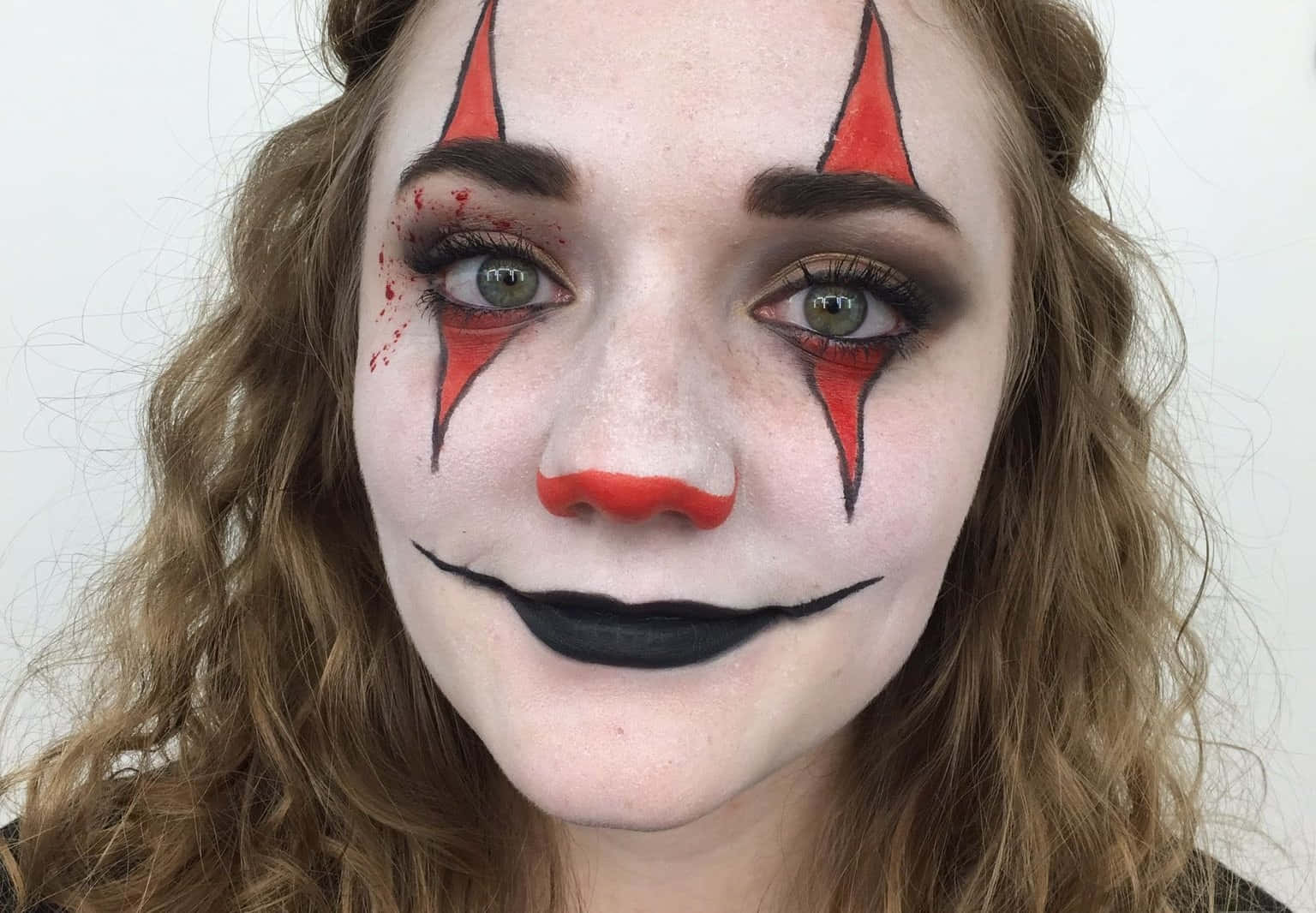 Halloween Face Paint 桌布 壁紙 — 一個人在純色背景上，帶有戲劇性的小丑妝容，包括紅色三角形、黑色嘴唇和類似血跡的污漬。