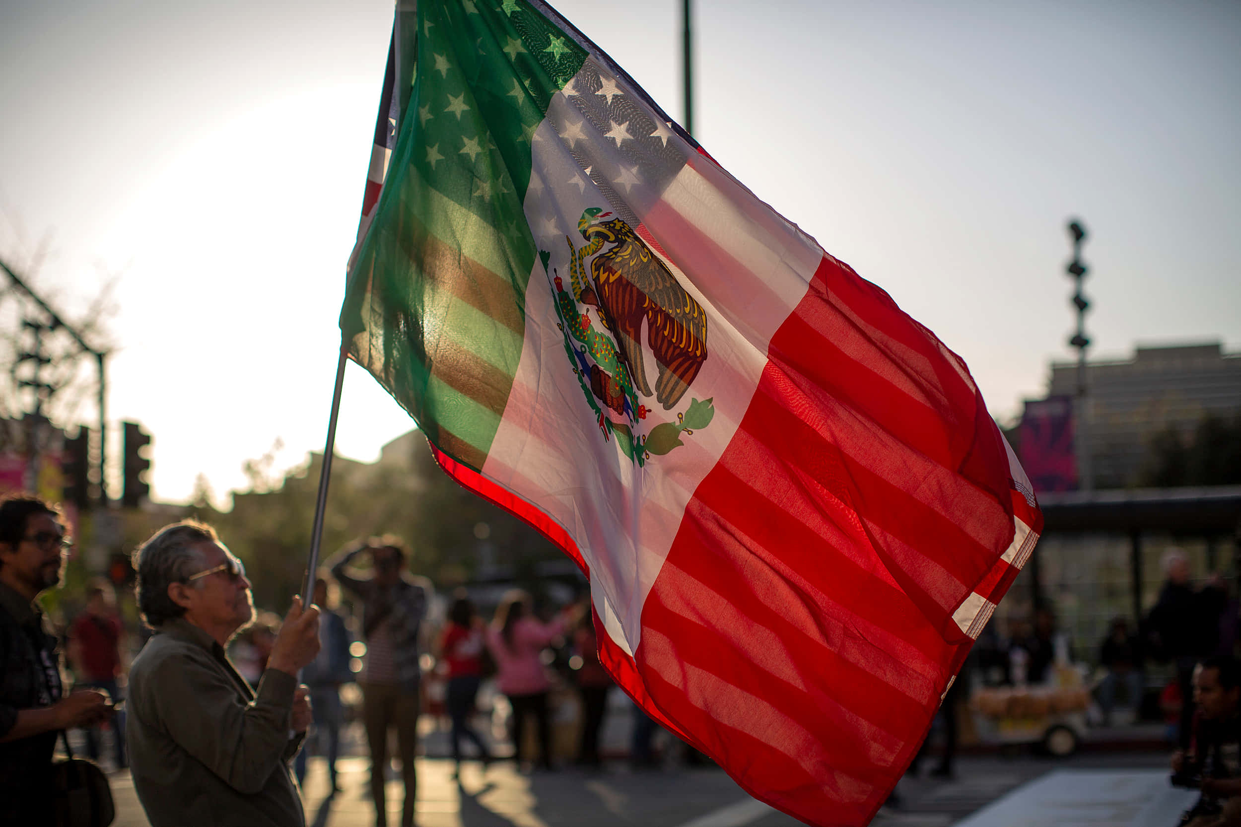 Mexican 桌布 壁紙 — 一個人在繁忙的城市街道上，手持一面在風中飄揚的墨西哥國旗，背景是明亮的日落。