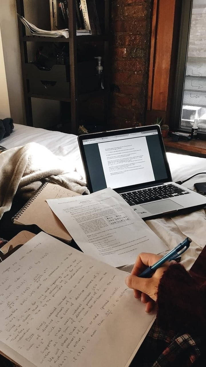Study Aesthetic 桌布 壁紙 — 一個人在舒適的陽光臥室裡，一邊在筆記型電腦上工作，一邊手寫筆記，房間有裸露磚牆和窗景。