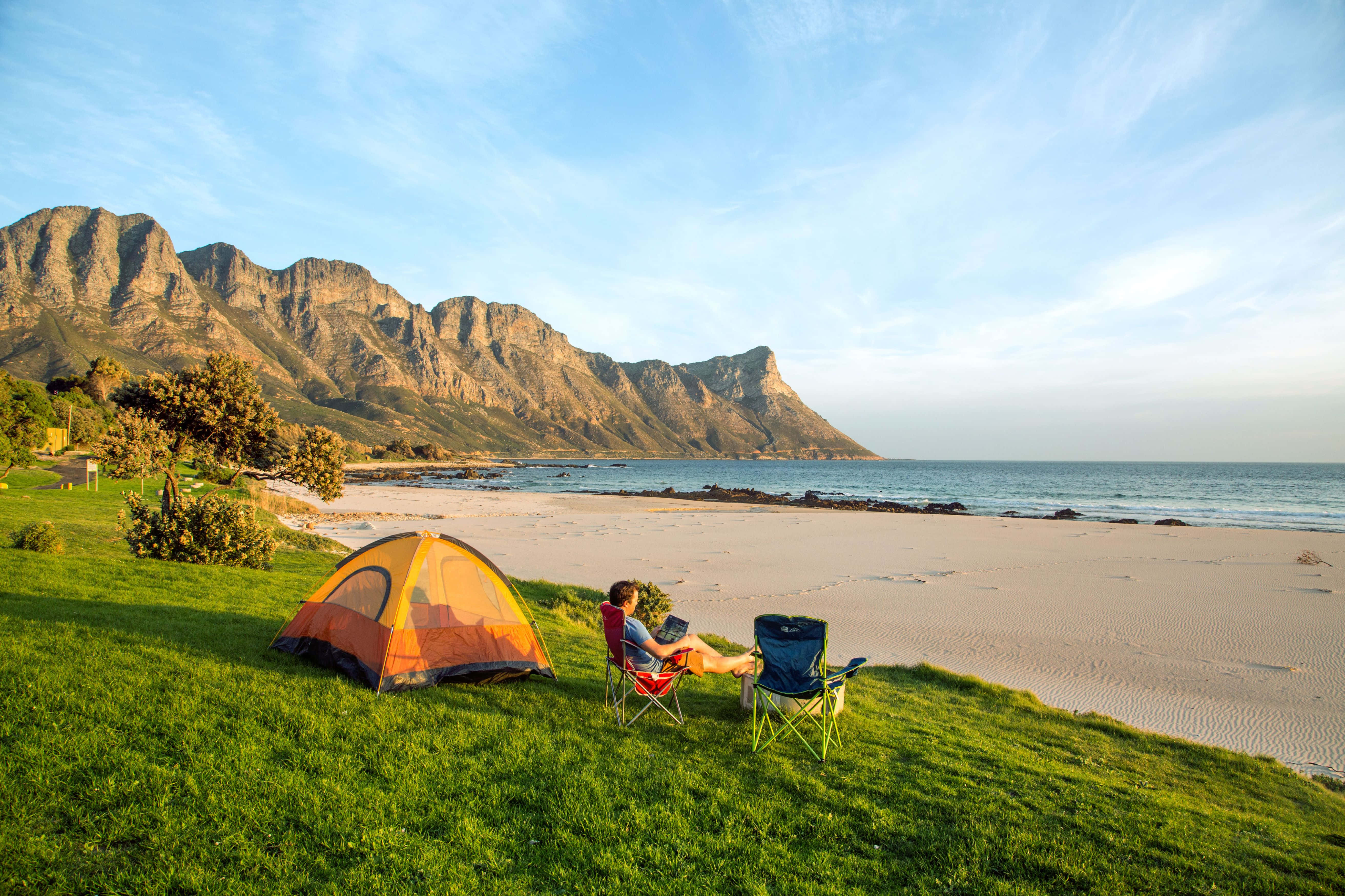 Beach Camping 桌布 壁紙 — 一個人在草地上嘅山丘上，喺橙色帳篷旁邊嘅椅子上放鬆，俯瞰沙灘同高聳嘅山脈，天空晴朗湛藍。