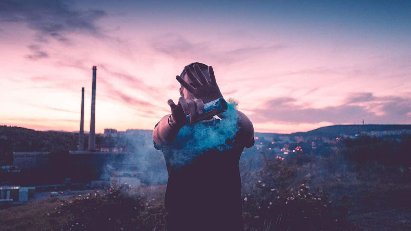 Chill 桌布 壁紙 — 一個人在黃昏時份戶外，從罐中釋放藍色煙霧，背景是粉紅和紫色的天空，遠處有工業煙囪。