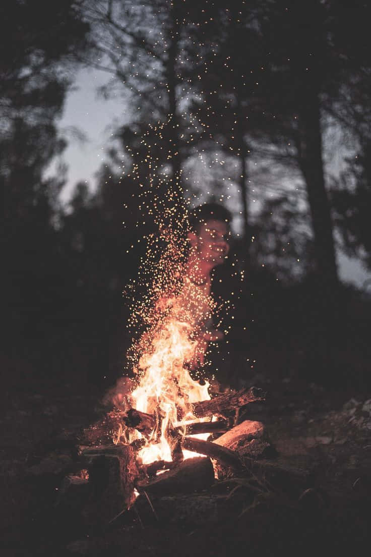 Campfire 桌布 壁紙 — 一個人在黃昏時分的黑暗森林中，以熊熊營火為背景，剪影中升起發光的餘燼。
