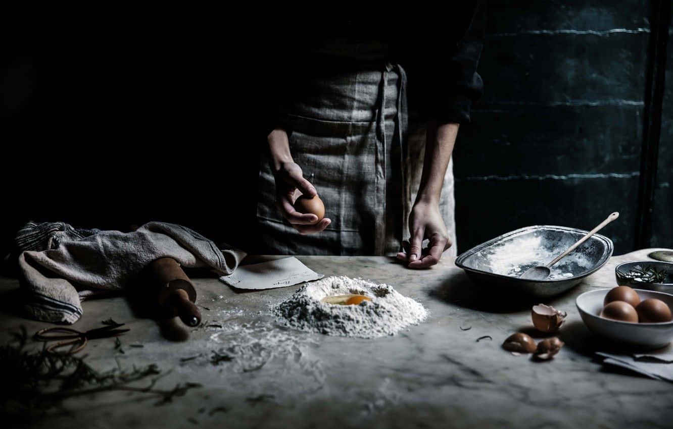 Cooking 桌布 壁紙 — 一個人在黑暗的廚房裡，將雞蛋打入石檯上的麵粉堆中，周圍擺放著烘焙工具。