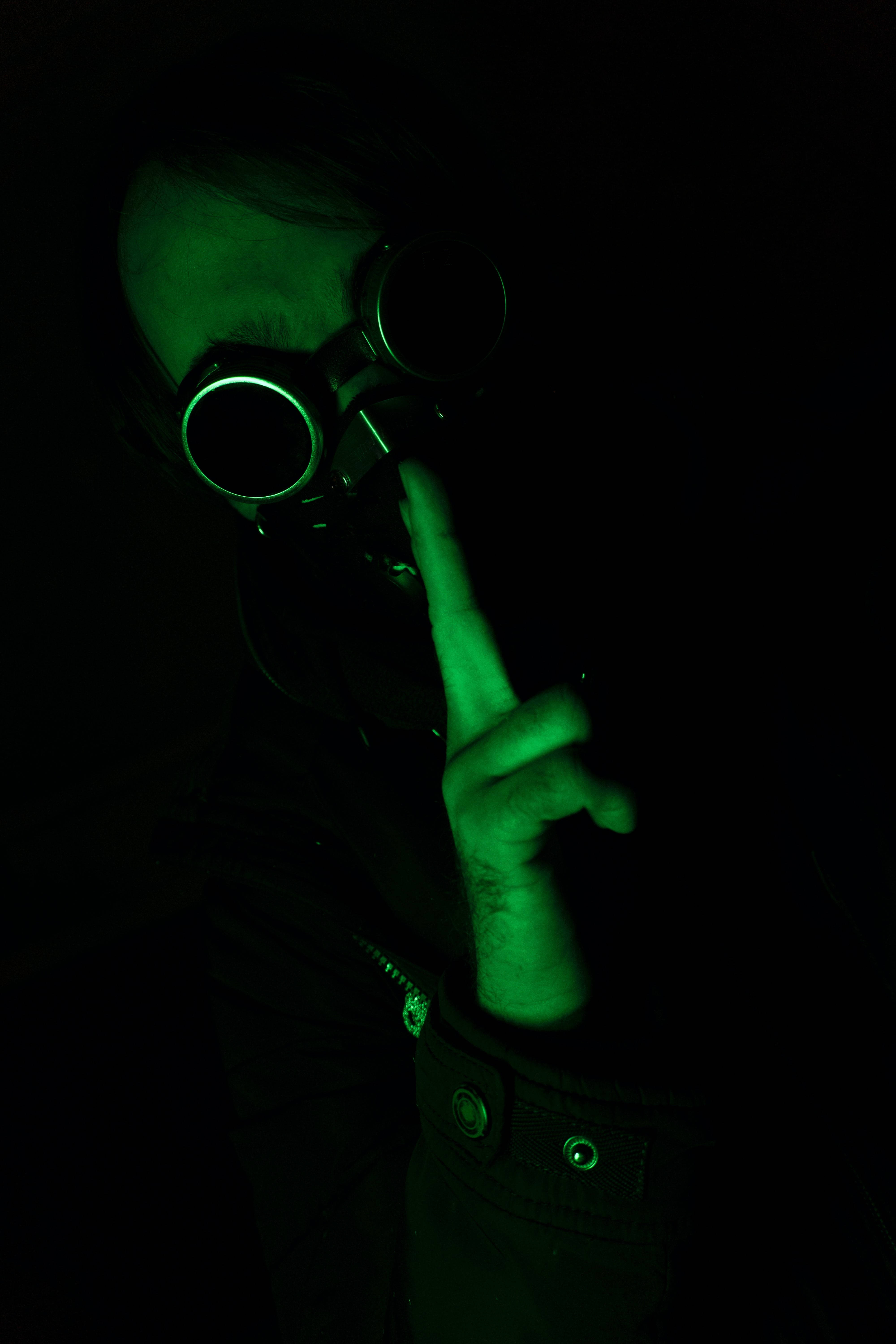 Black And Green 桌布 壁紙 — 一個人在黑暗的房間中被詭異的綠光照亮，戴著護目鏡並將手指放在嘴唇上示意安靜。
