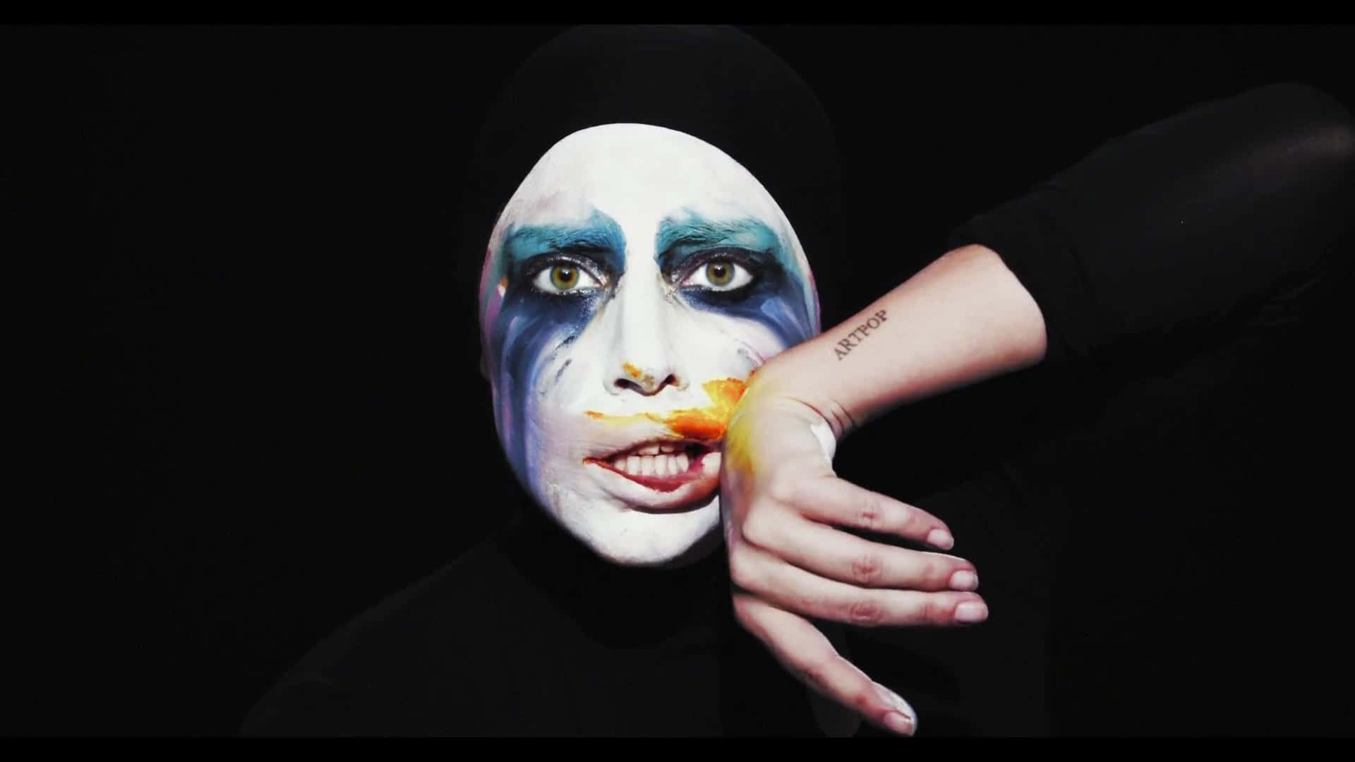 Lady Gaga 桌布 壁紙 — 一個人在黑色背景前，臉上帶有戲劇性的藍白彩繪和橙色唇紋，手靠近臉部。