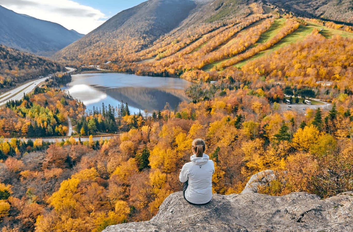 Fall Hikes 桌布 壁紙 — 一個人坐在岩石上，俯瞰著被鮮豔秋葉和起伏山丘環繞的寧靜湖泊。