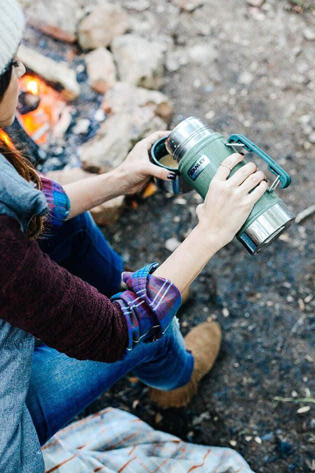 Minimalist Camping 桌布 壁紙 — 一個人坐在營火旁，手持綠色保溫瓶，戴著冷帽和格仔恤衫。