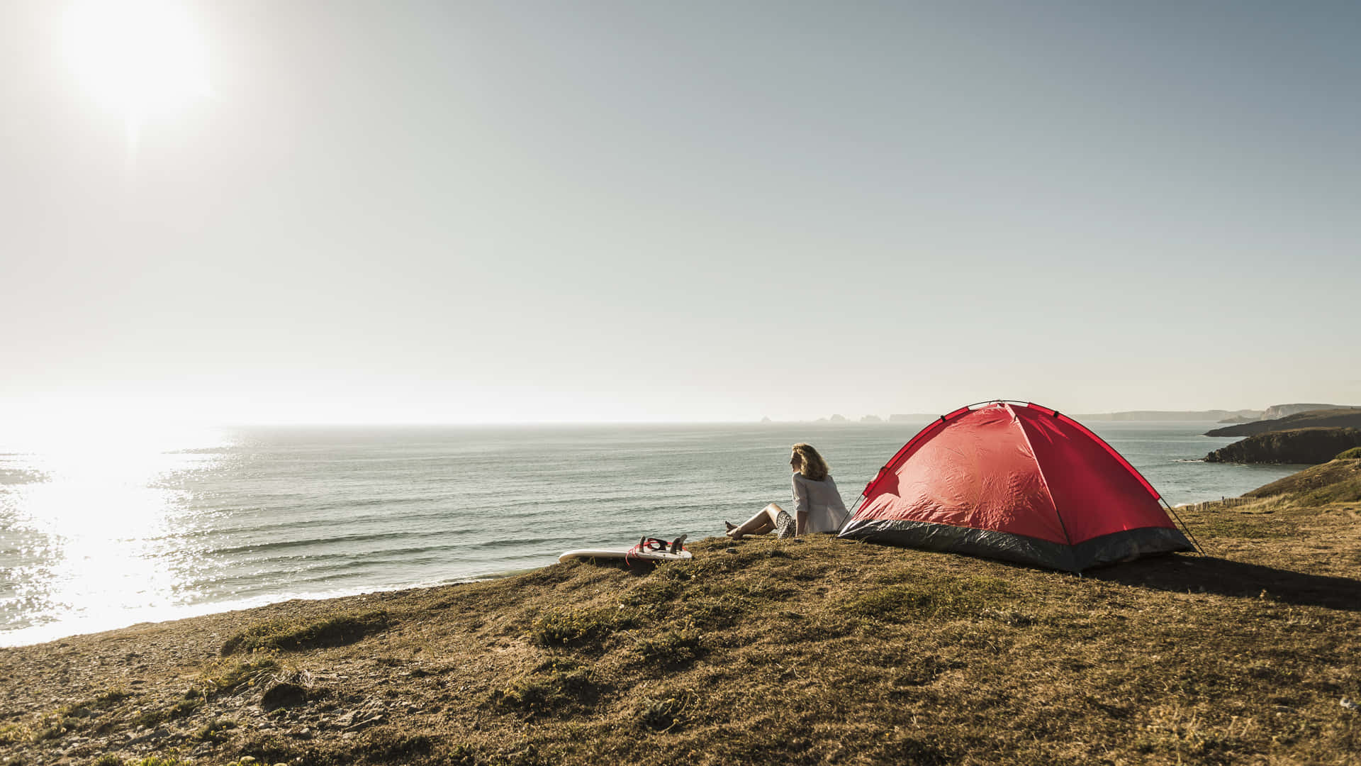 Beach Camping 桌布 壁紙 — 一個人坐在草地上嘅懸崖邊，旁邊有個紅色帳篷，俯瞰陽光普照嘅海洋同遠處島嶼。