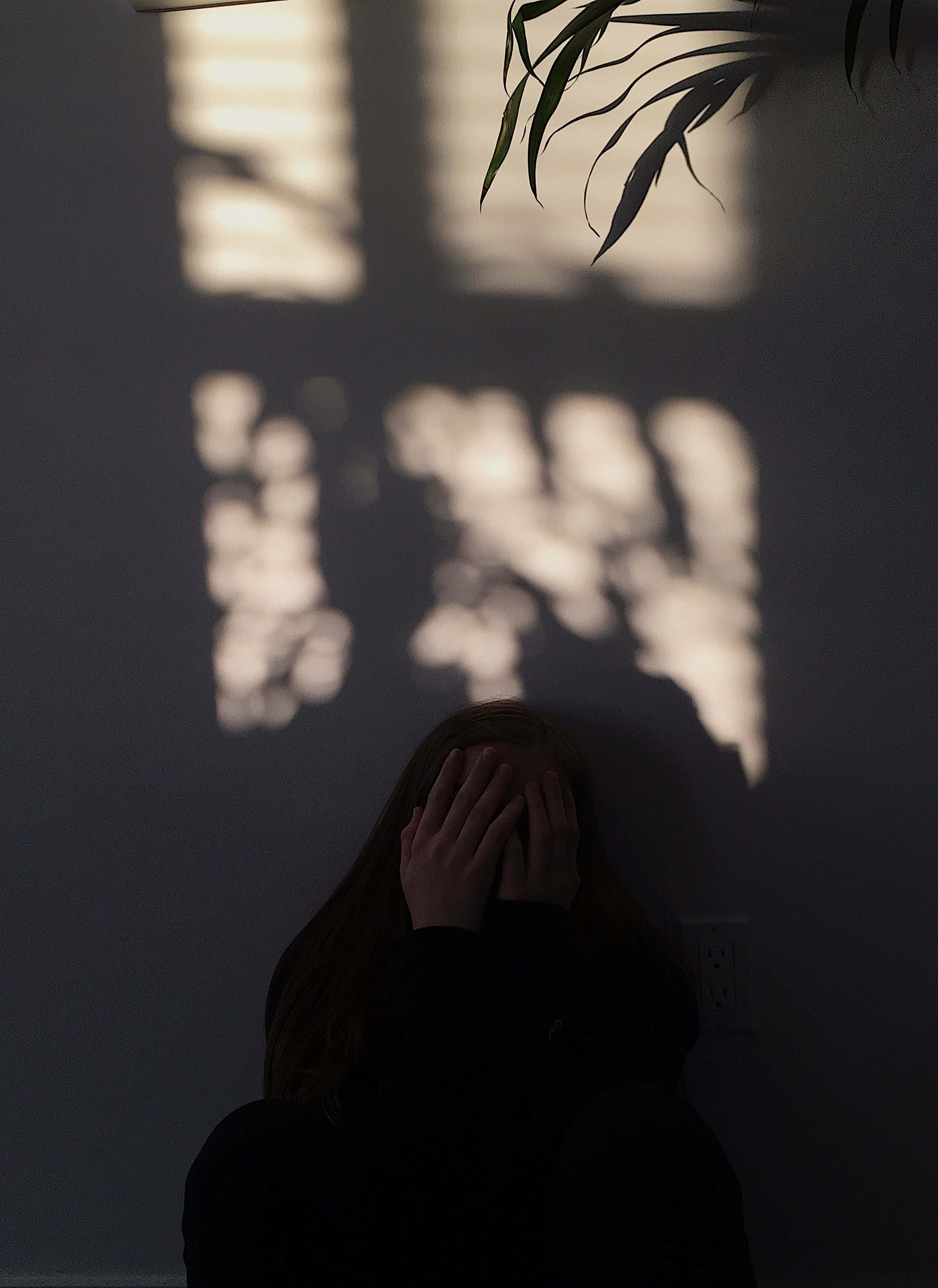 sadness 桌布 壁紙 — 一個人坐在陰影中，雙手掩面，柔和陽光在他們身後的牆上投射出葉狀圖案。