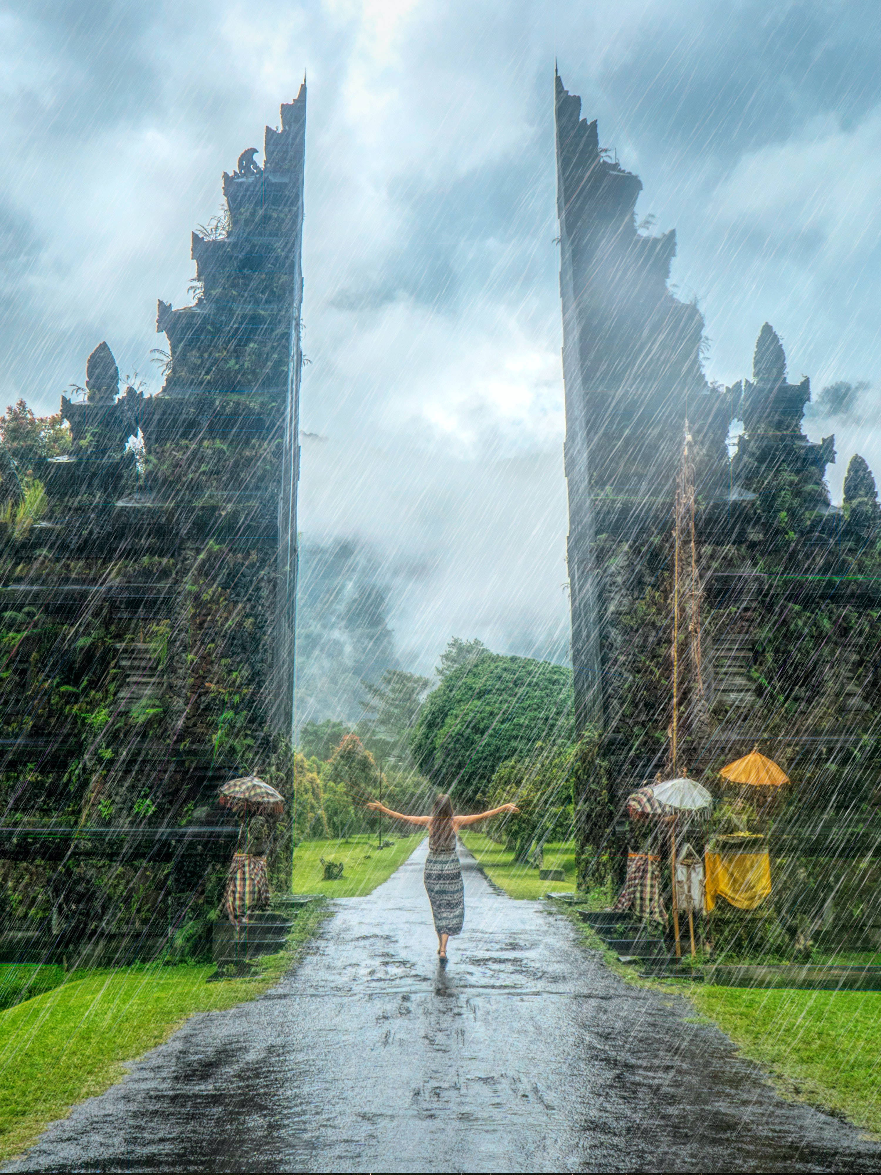 Rainy 桌布 壁紙 — 一個人張開雙臂，在風暴天空下，沿著一條濕滑的雨路，走在兩座高聳的古老石門之間。