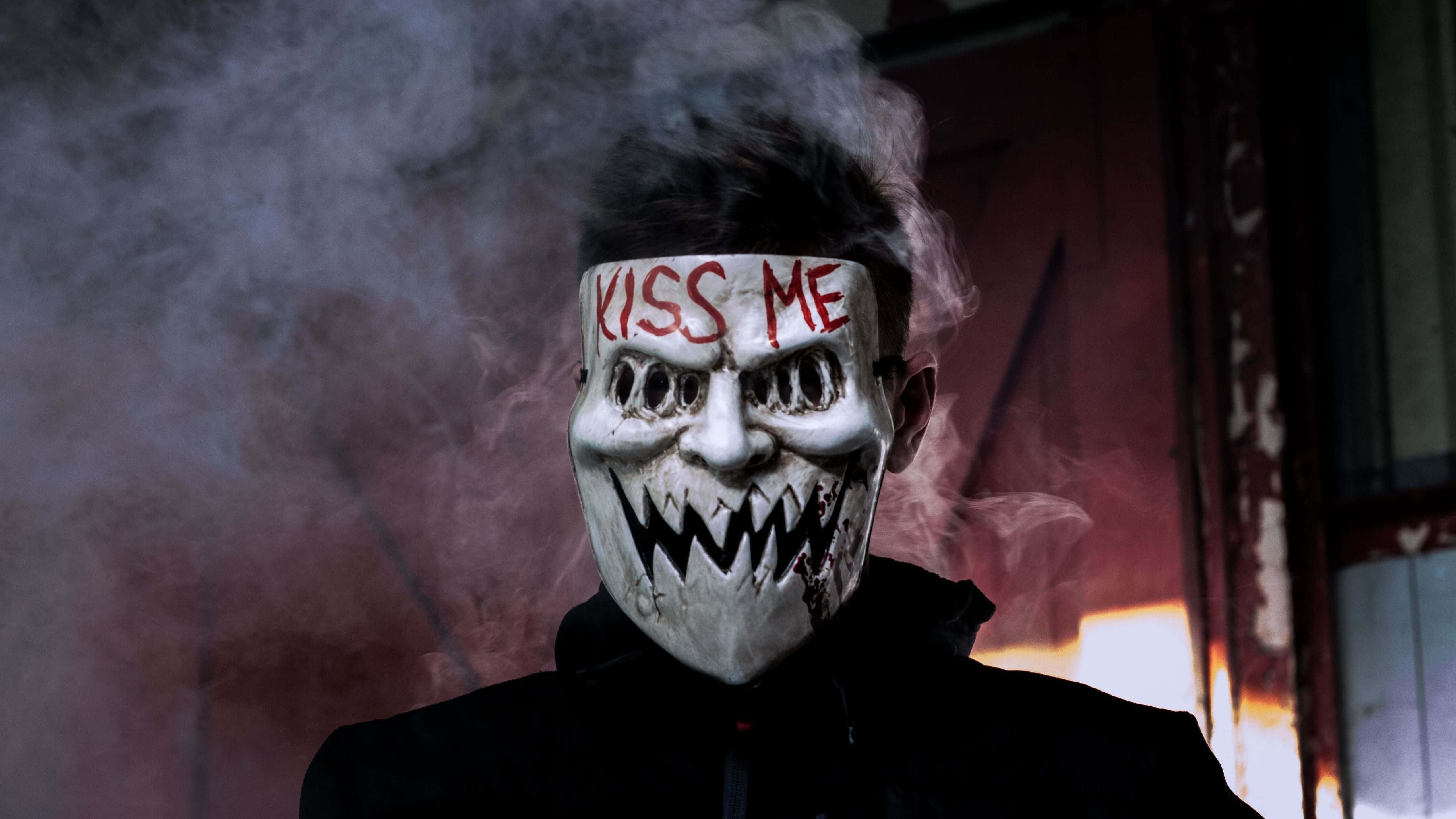 Horror Masks 桌布 壁紙 — 一個人戴住白色恐怖面具，上面有紅色「KISS ME」字樣同鋸齒狀牙齒，企喺煙霧瀰漫嘅昏暗光線中，靠近一個燃燒緊嘅門口。