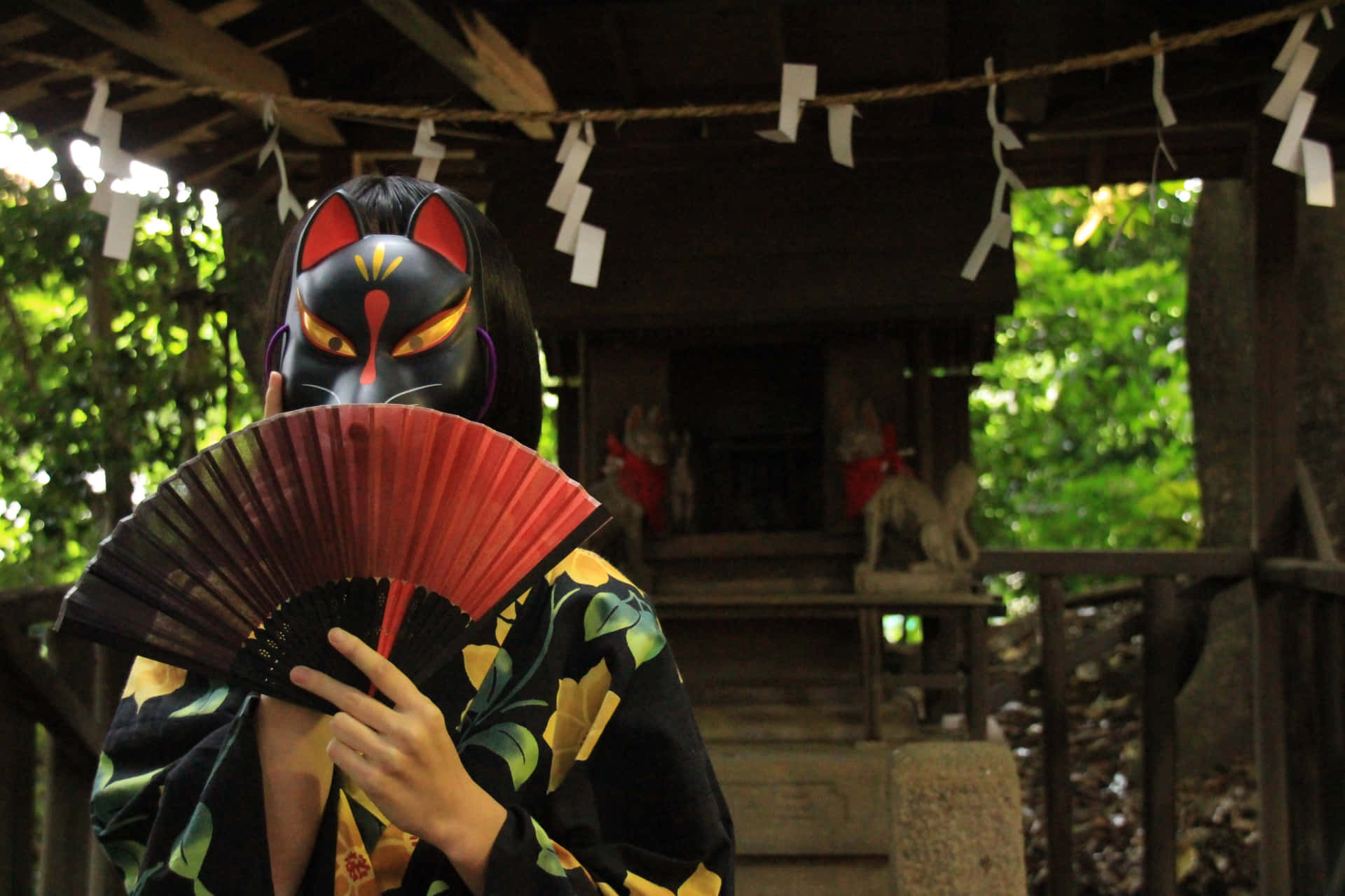 Tengu 桌布 壁紙 — 一個人戴住黑色狐狸面具、著住花卉和服，喺木造神社拎住紅色扇子，周圍係茂密嘅綠葉。