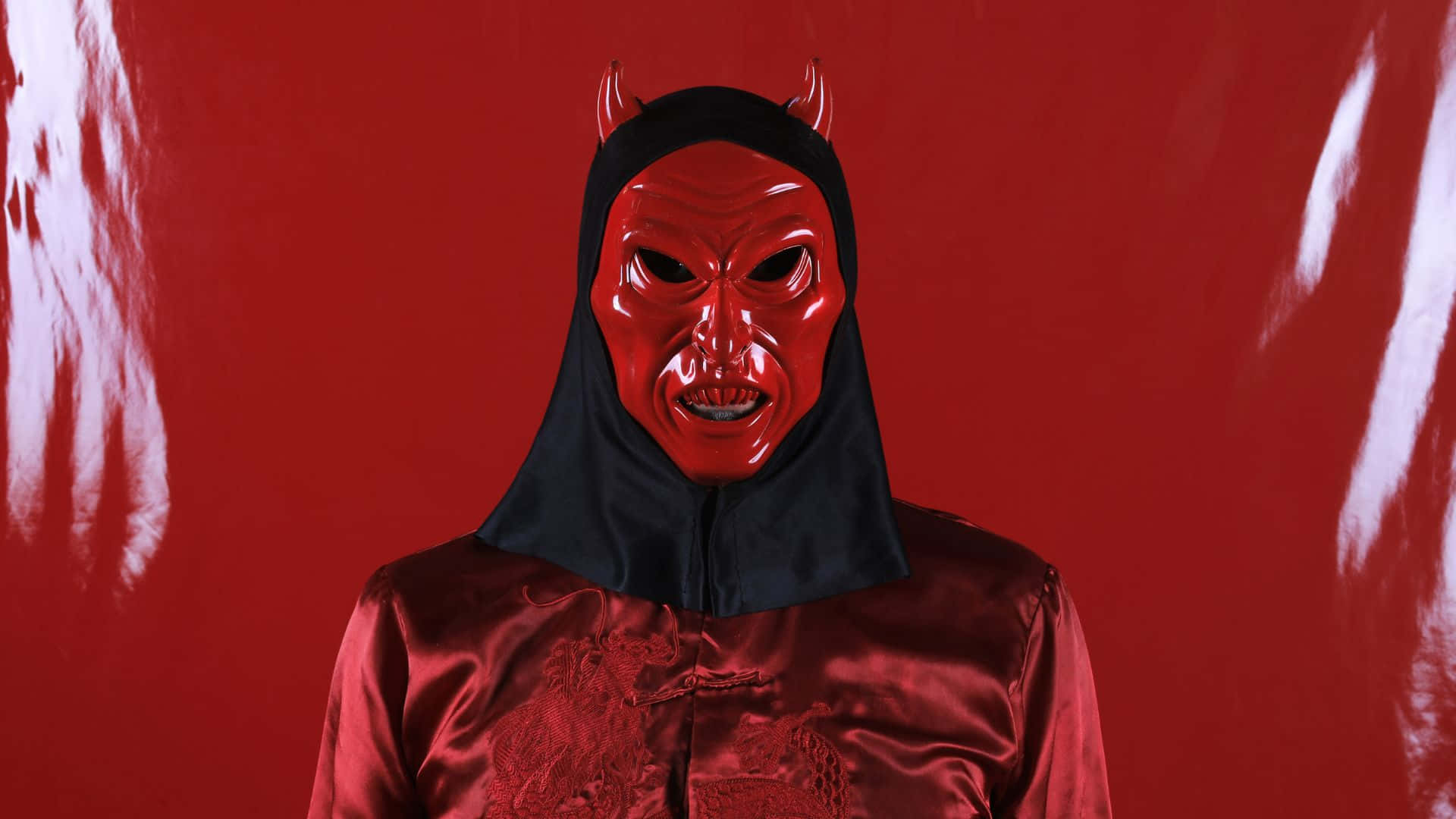 Horror Masks 桌布 壁紙 — 一個人戴著光面紅色魔鬼面具，有角及黑色頭罩，站在有白色油漆條紋的鮮紅色背景前。