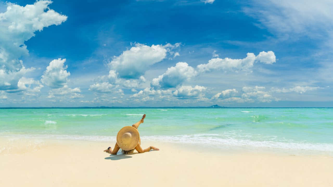 Jamaican Beach 桌布 壁紙 — 一個人戴著寬邊帽躺在純淨的白色沙灘上，沙灘延伸至碧綠的海水，上方是廣闊的藍天和蓬鬆的白雲。