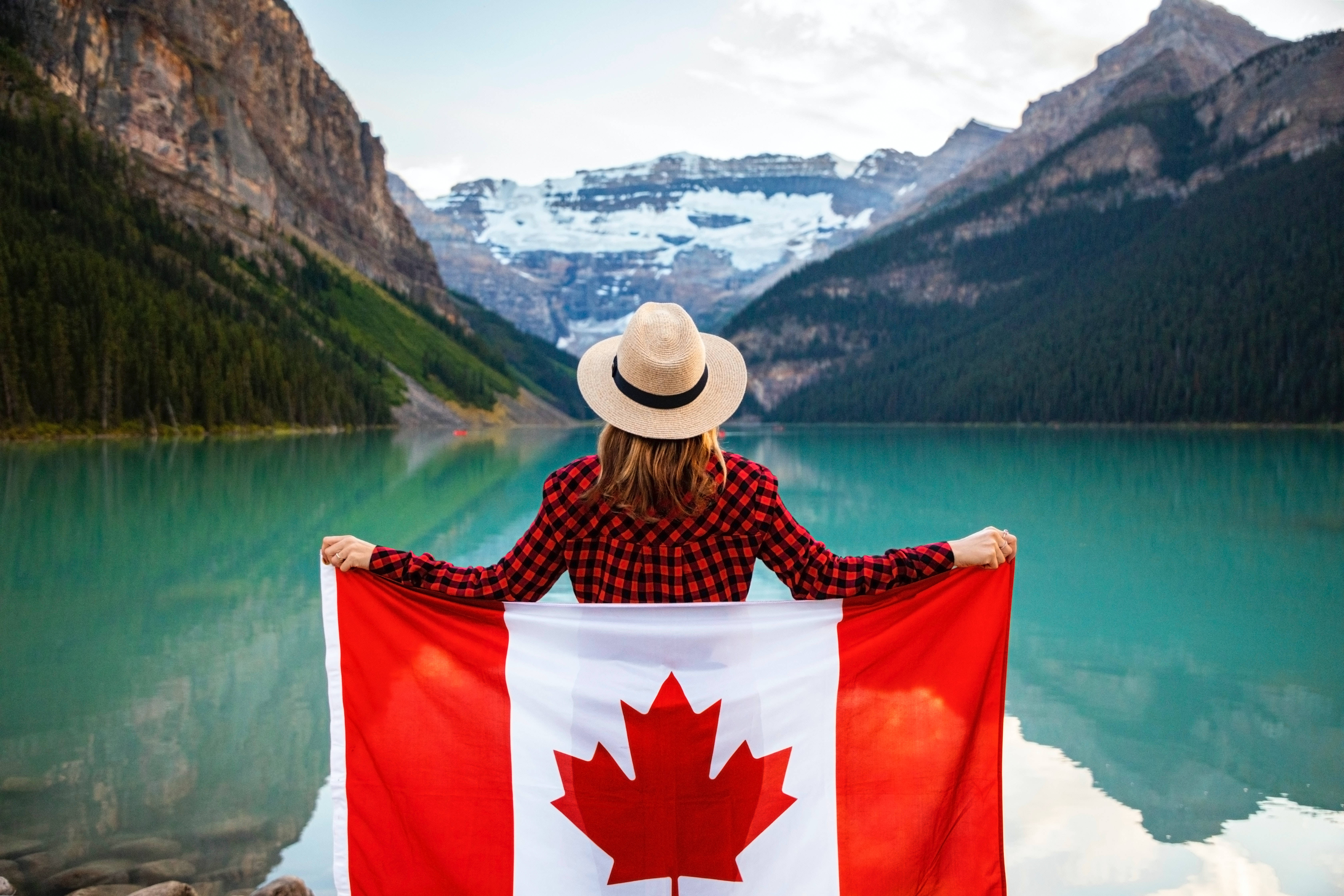 Canadian 桌布 壁紙 — 一個人戴著帽子和穿著格仔恤衫，在一個平靜水面倒映著雪峰的藍綠色山湖邊舉起加拿大國旗。