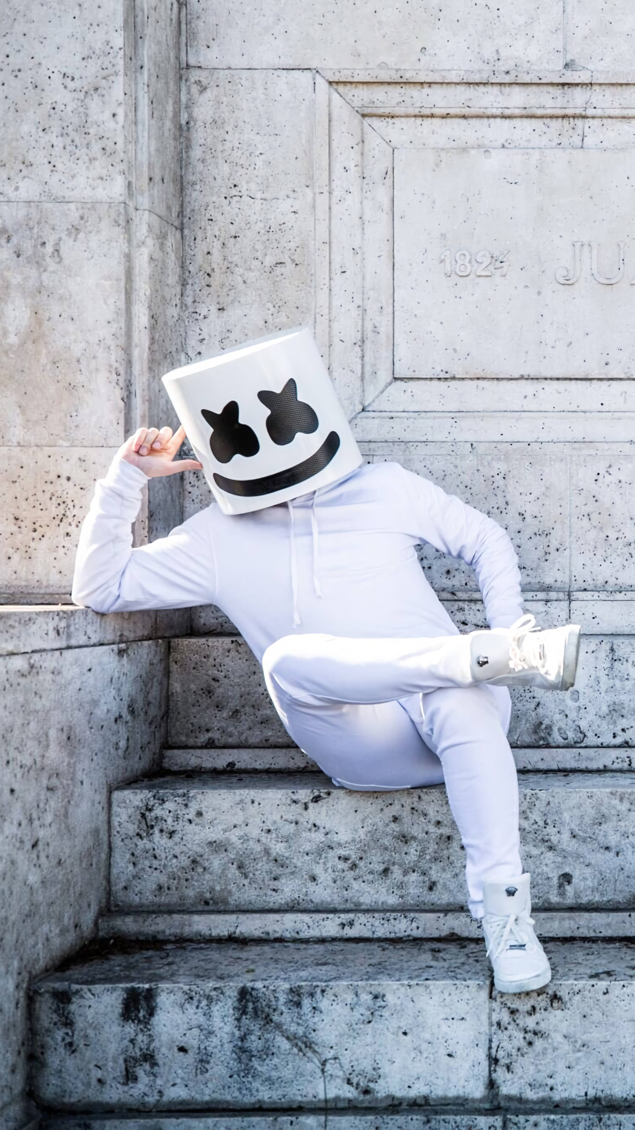 marshmello iPhone 桌布 壁紙 — 一個人戴著白色棉花糖面具，身穿全白服裝，隨意地坐在風化的石階上，背景是帶有紋理的牆壁。