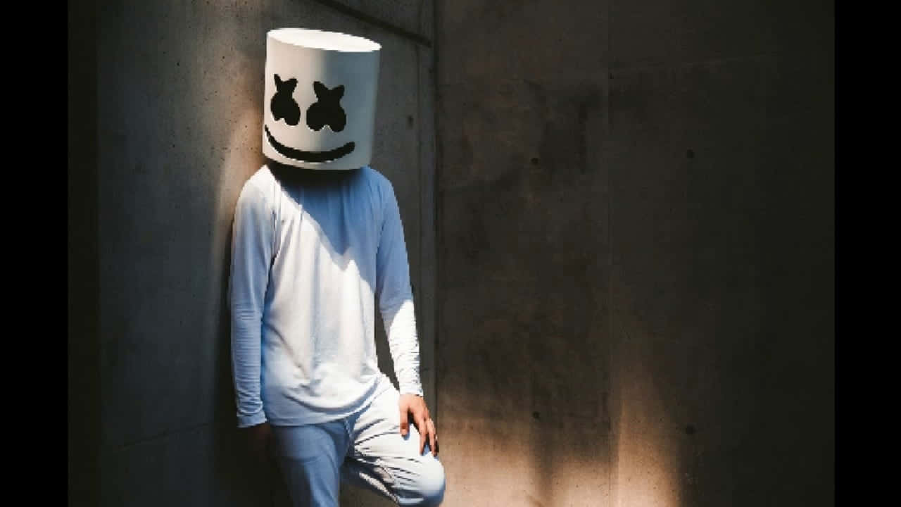 Marshmello 桌布 壁紙 — 一個人戴著白色棉花糖頭盔，面帶微笑，靠著混凝土牆，沐浴在斑駁的陽光下。