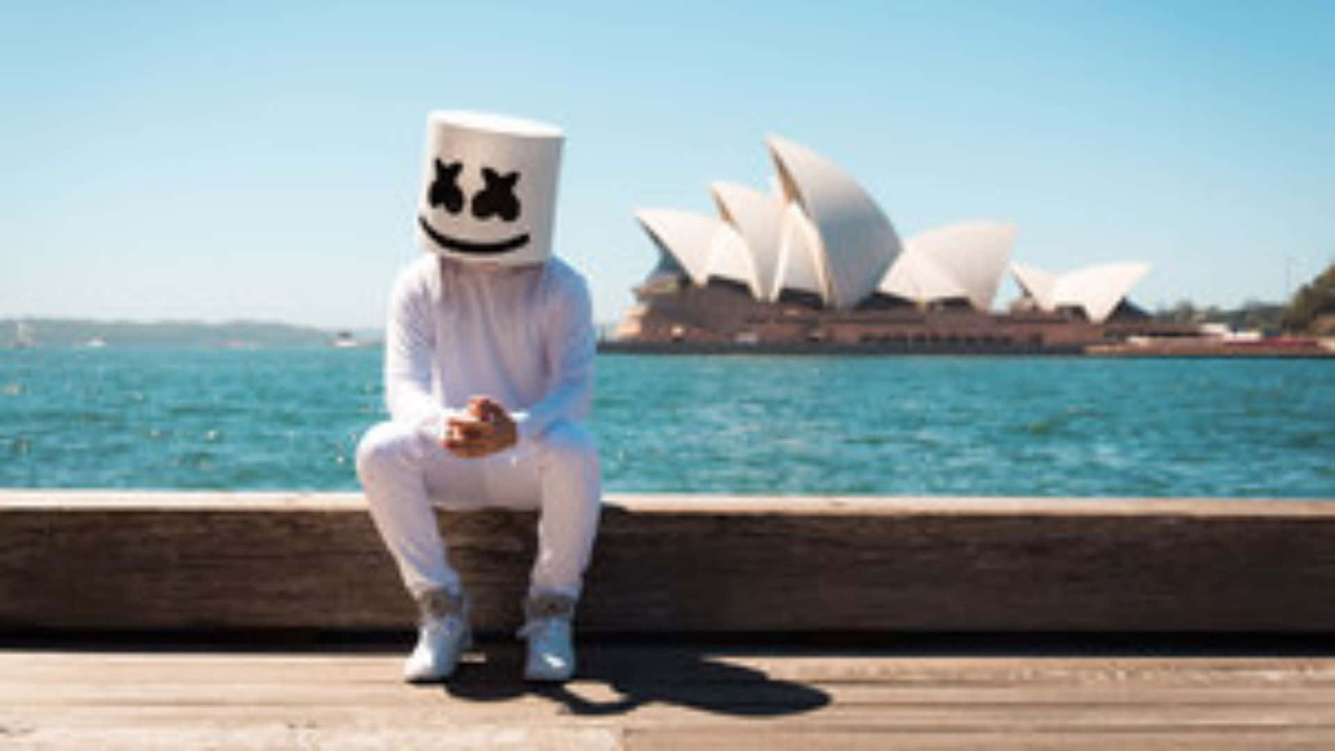 Marshmello 桌布 壁紙 — 一個人戴著白色棉花糖頭盔坐在木碼頭上，背景是悉尼歌劇院，天空清澈湛藍。