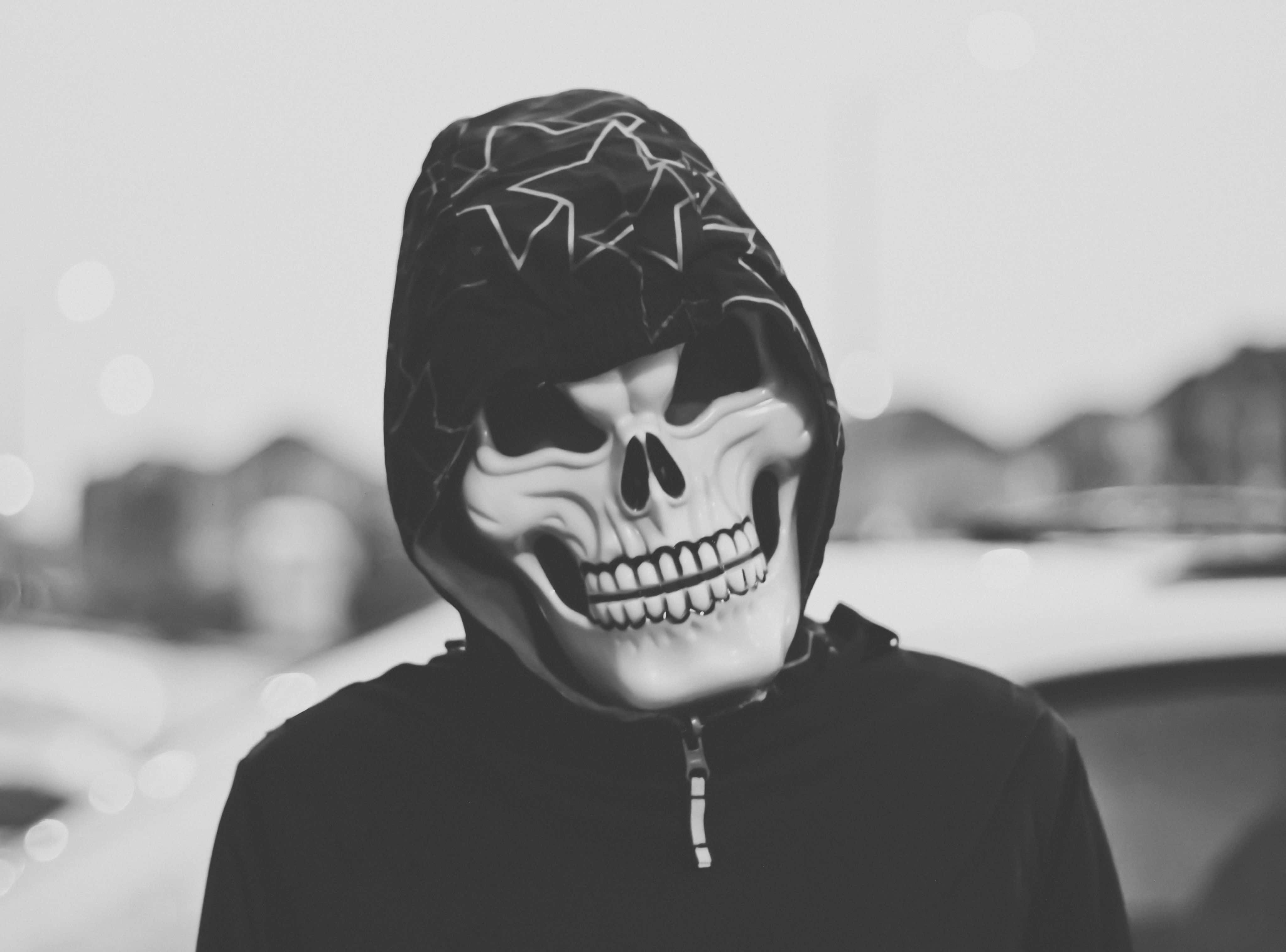 Horror Masks 桌布 壁紙 — 一個人戴著骷髏面具和連帽外套，身處模糊的城市環境中，背景有散景燈光。