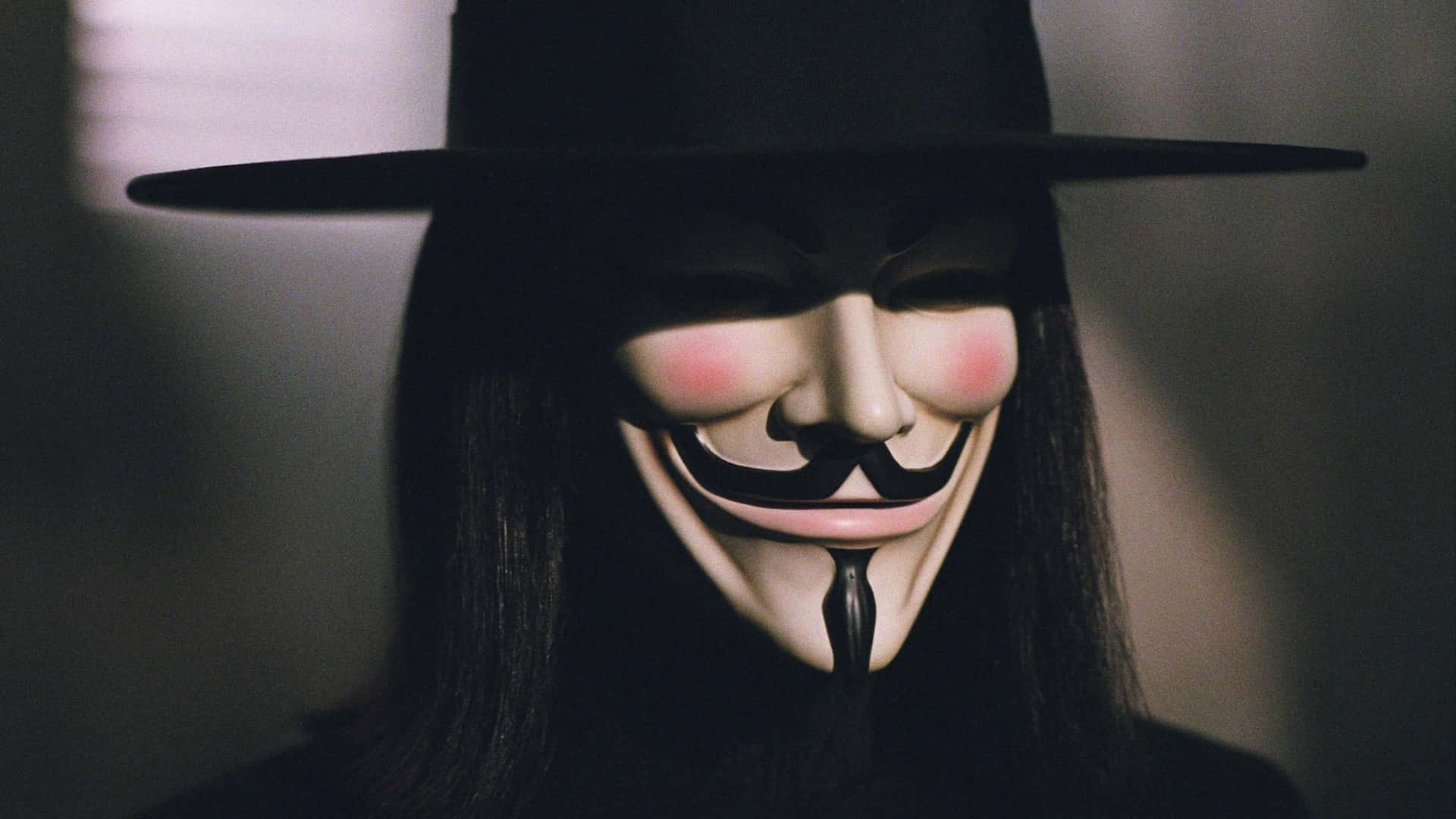 Halloween Masks 桌布 壁紙 — 一個人戴著黑色帽子和標誌性的蓋伊·福克斯面具，臉頰紅潤，咧嘴大笑，站在昏暗的燈光下。