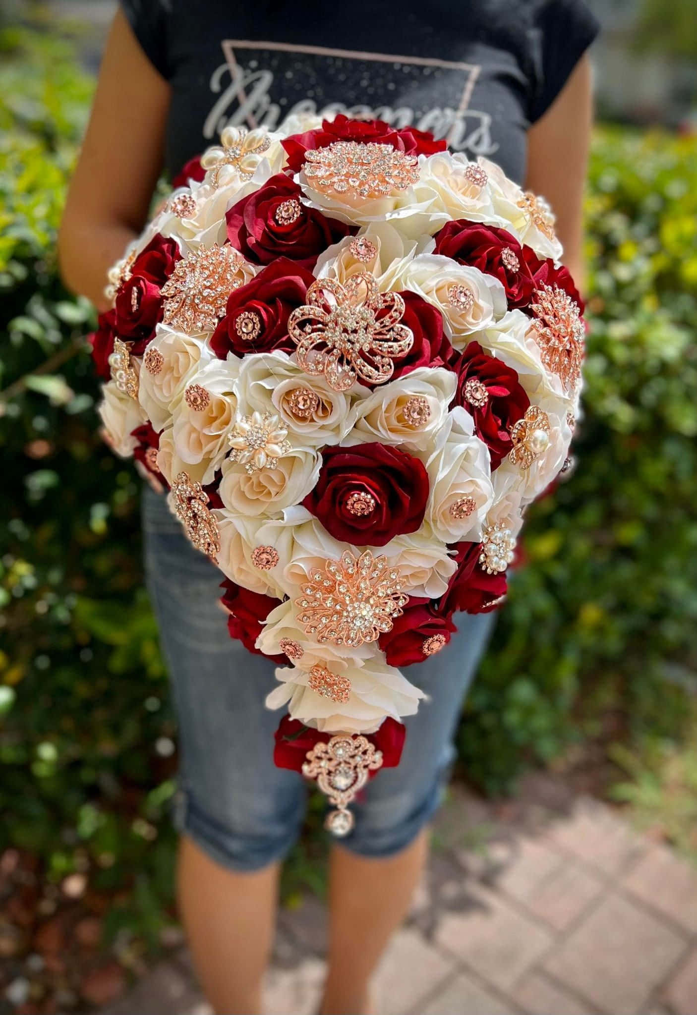 Roses Wedding 桌布 壁紙 — 一個人手持一束奢華的深紅和象牙色玫瑰瀑布花束，飾以閃閃發光的玫瑰金胸針，背景是鬱鬱蔥蔥的花園。