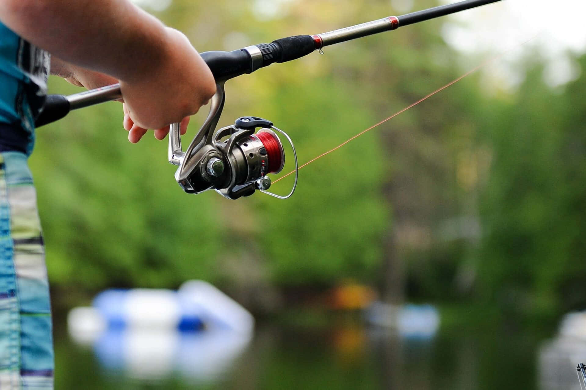 Fishing Rod 桌布 壁紙 — 一個人手緊握魚竿的特寫，魚竿帶有紅色線輪，背景是模糊的綠色森林和水面。
