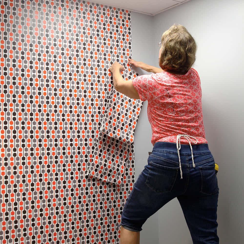 Installation Art 桌布 壁紙 — 一個人正在將帶有橙色、灰色和黑色幾何形狀的復古圖案壁紙貼在房間的素色牆壁上。