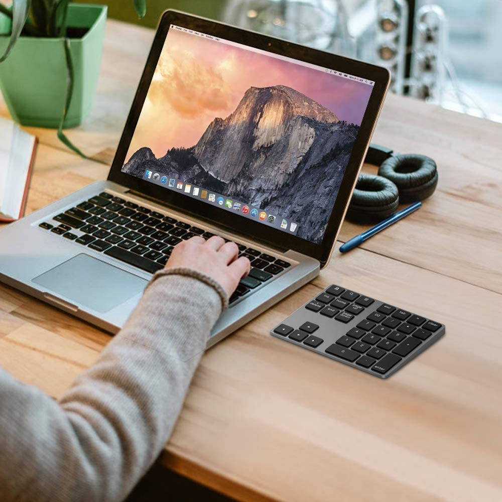 macbook 桌布 壁紙 — 一個人的手在銀色手提電腦上打字，電腦顯示山景日落桌布，旁邊有緊湊型鍵盤和耳機，放在木桌上。