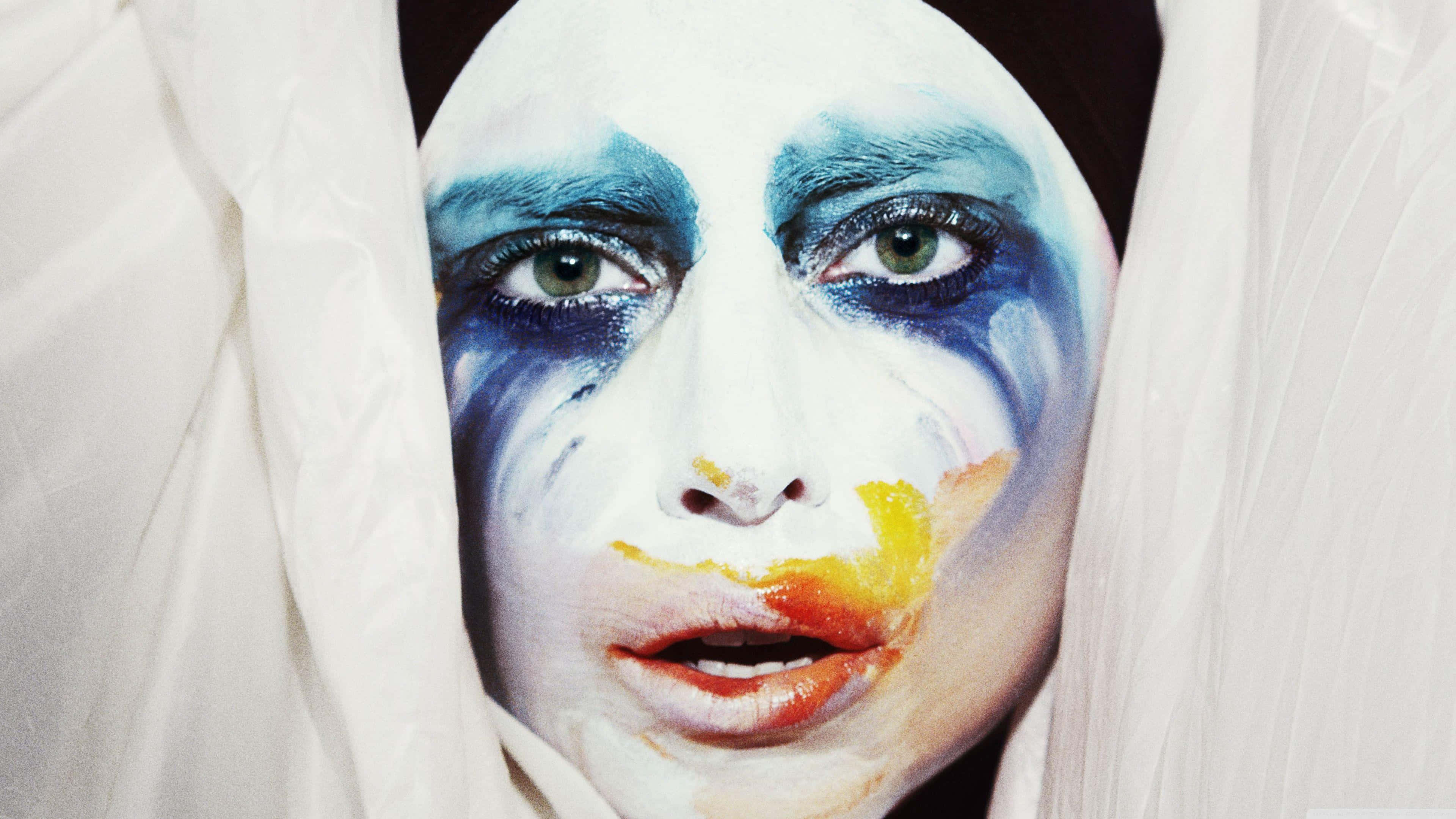 Lady Gaga 桌布 壁紙 — 一個人的特寫肖像，臉上帶有戲劇性的藍色和橙色顏料，從白色布料中窺視。