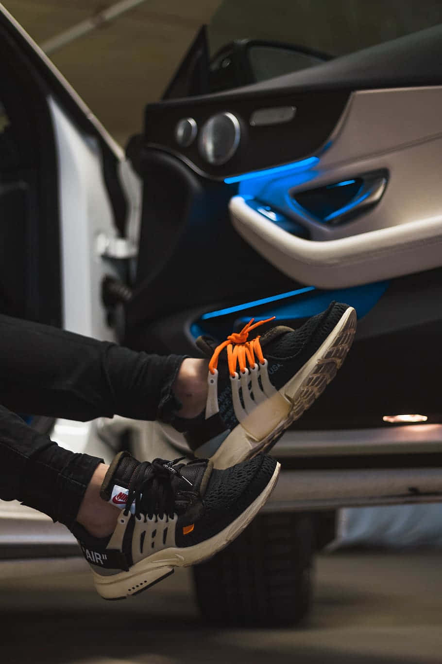 Footwear Design 桌布 壁紙 — 一個人的腳部特寫，穿著黑色運動鞋配橙色鞋帶，靠近打開的車門，車內有藍色燈光。