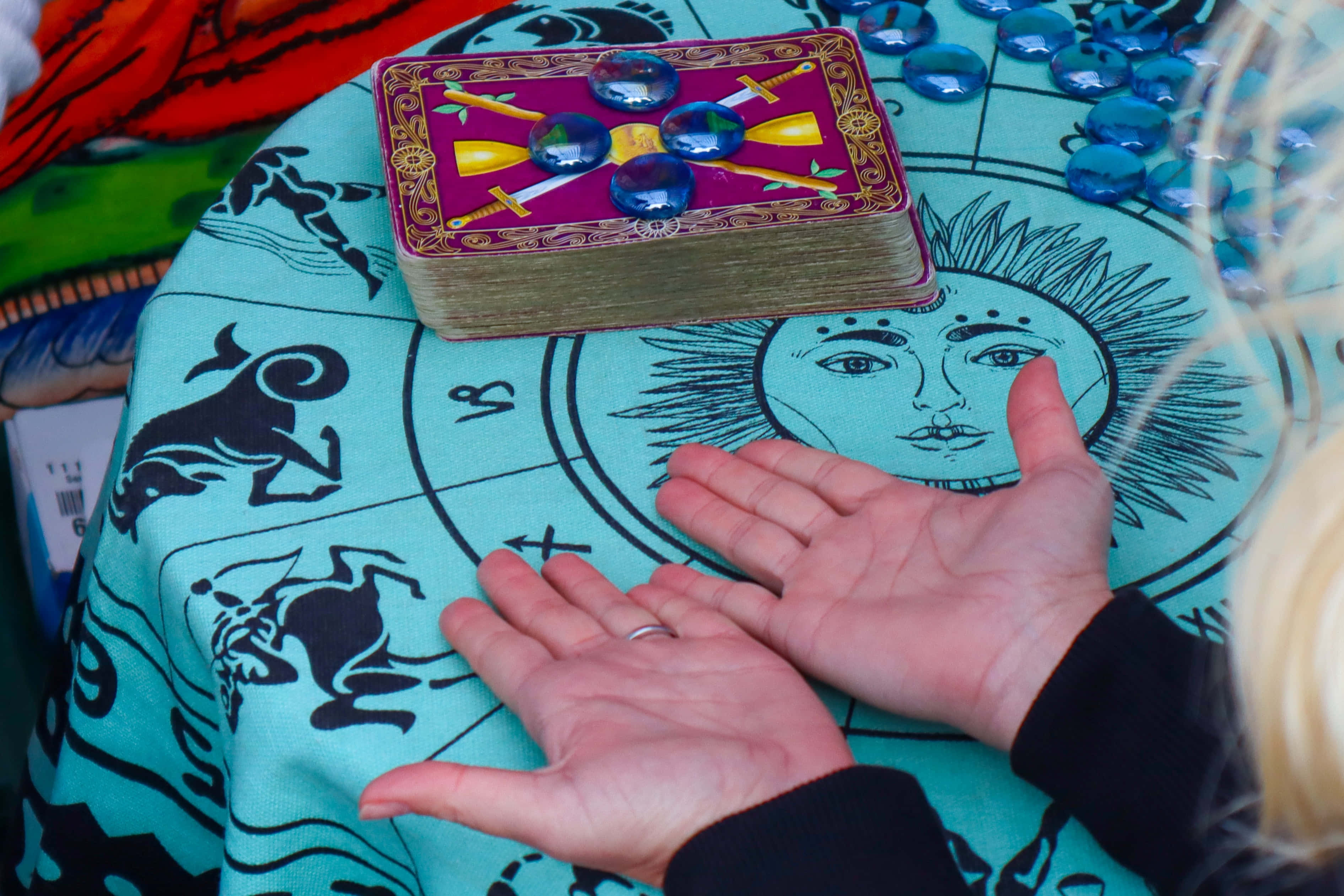 Tarot Card 桌布 壁紙 — 一個人的雙手張開，放在一塊飾有星座符號和太陽臉的神秘青綠色布上，附近有一副帶有藍色石頭的塔羅牌。