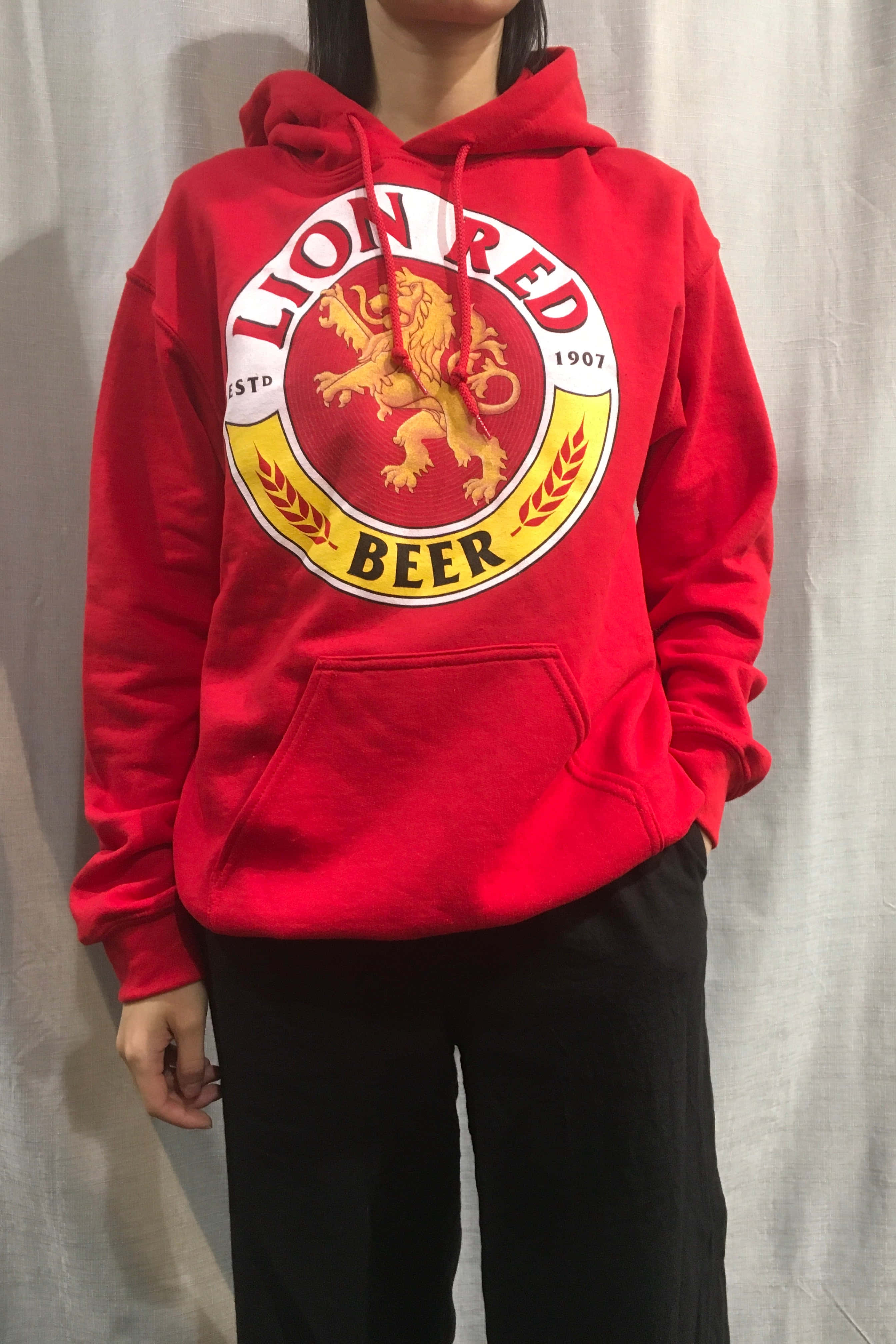 Red Hoodie 桌布 壁紙 — 一個人穿著一件大膽的紅色連帽衫，上面有大型Lion Red Beer標誌和金色獅子徽章，背景是純白色窗簾。