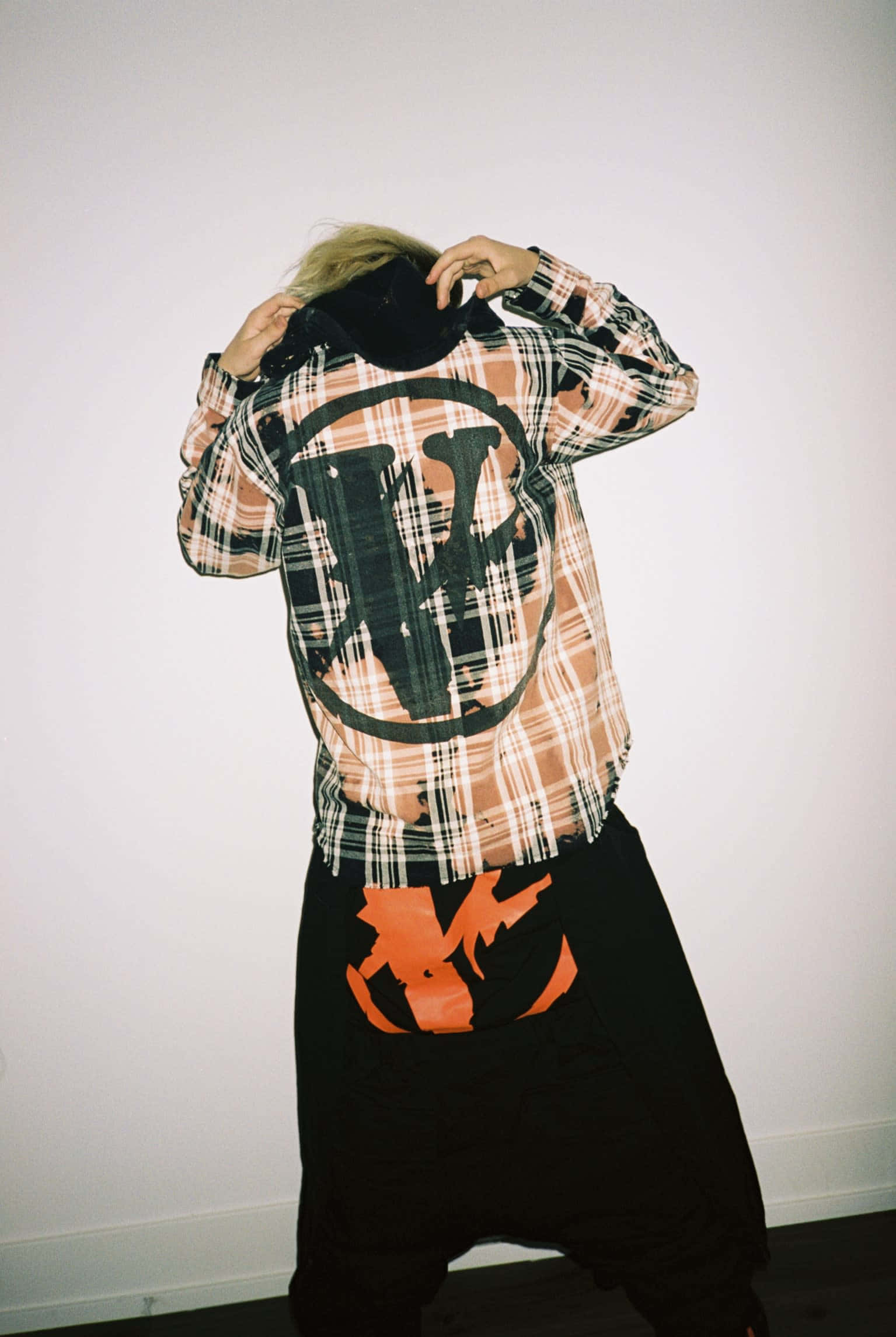 Vlone 桌布 壁紙 — 一個人穿著格仔衫，上面有大型黑色標誌和磨邊下擺，轉身背對，雙手調整帽子，背景是純白牆壁。