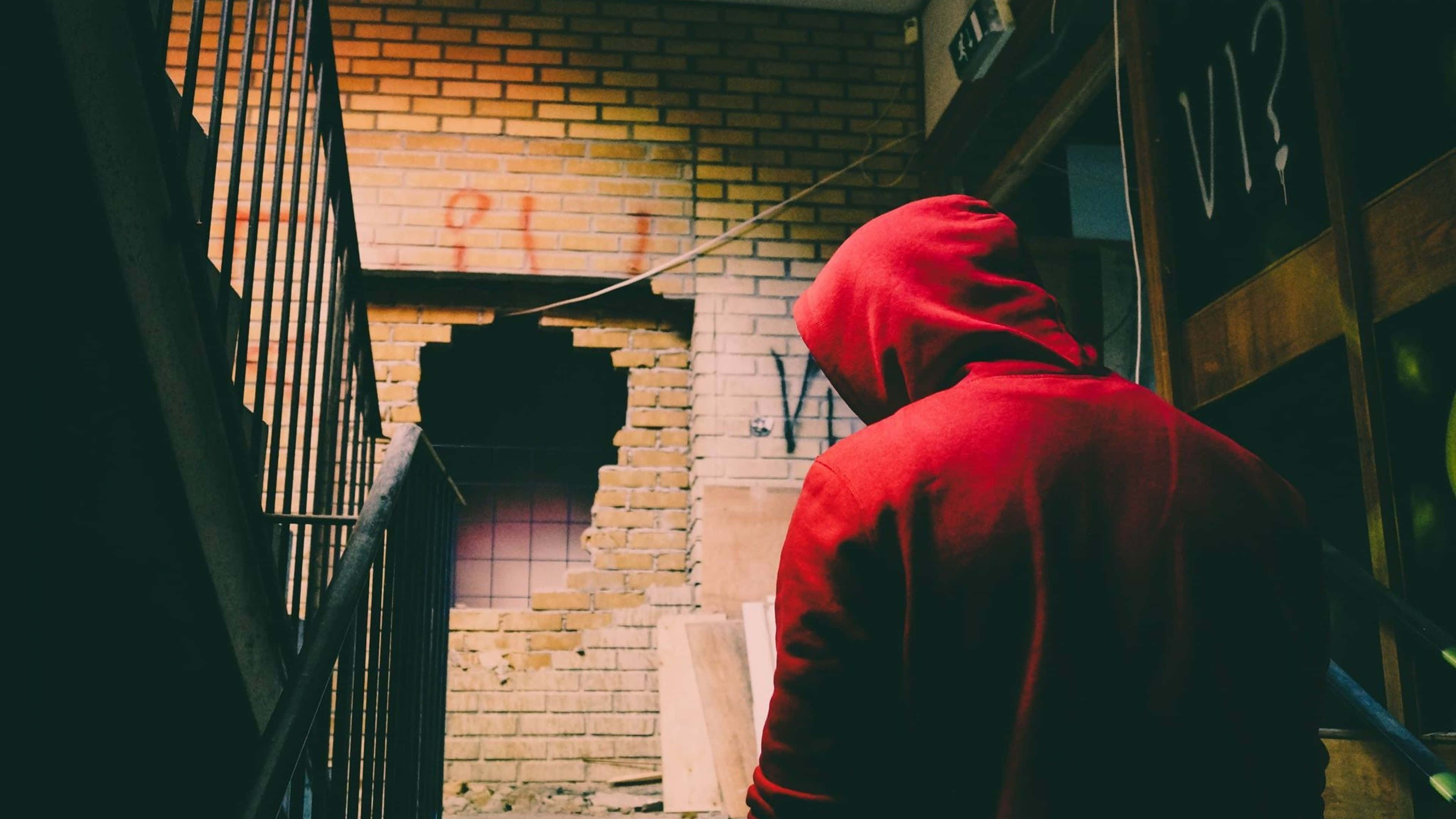 Red Hoodie 桌布 壁紙 — 一個人穿著紅色連帽衫，站在一條小巷裡，旁邊是破損的磚牆，牆上有塗鴉，在溫暖的環境光線下。