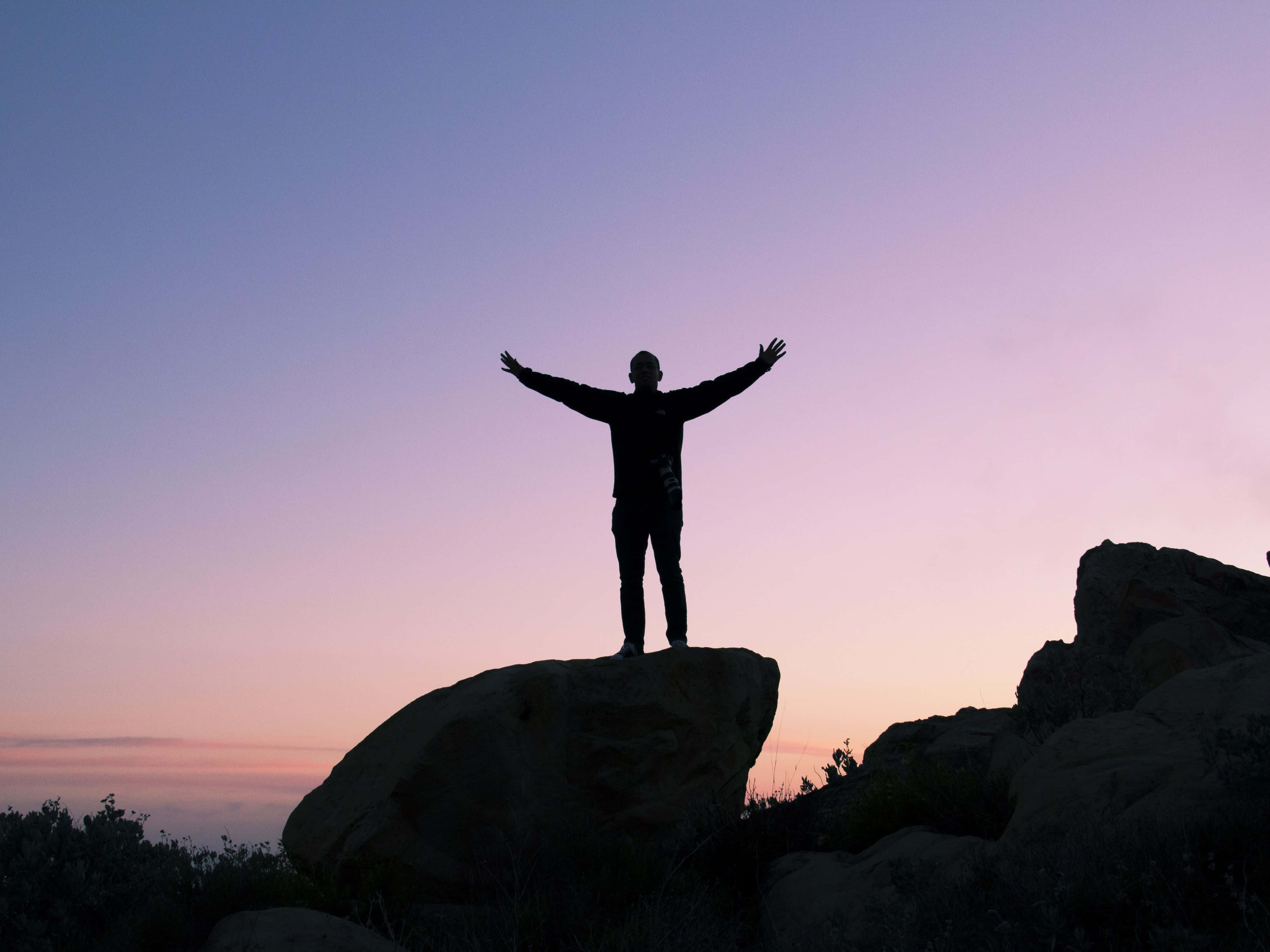 Success 桌布 壁紙 — 一個人站在岩石上，雙臂張開，背景是柔和的紫色和粉紅色暮色天空。