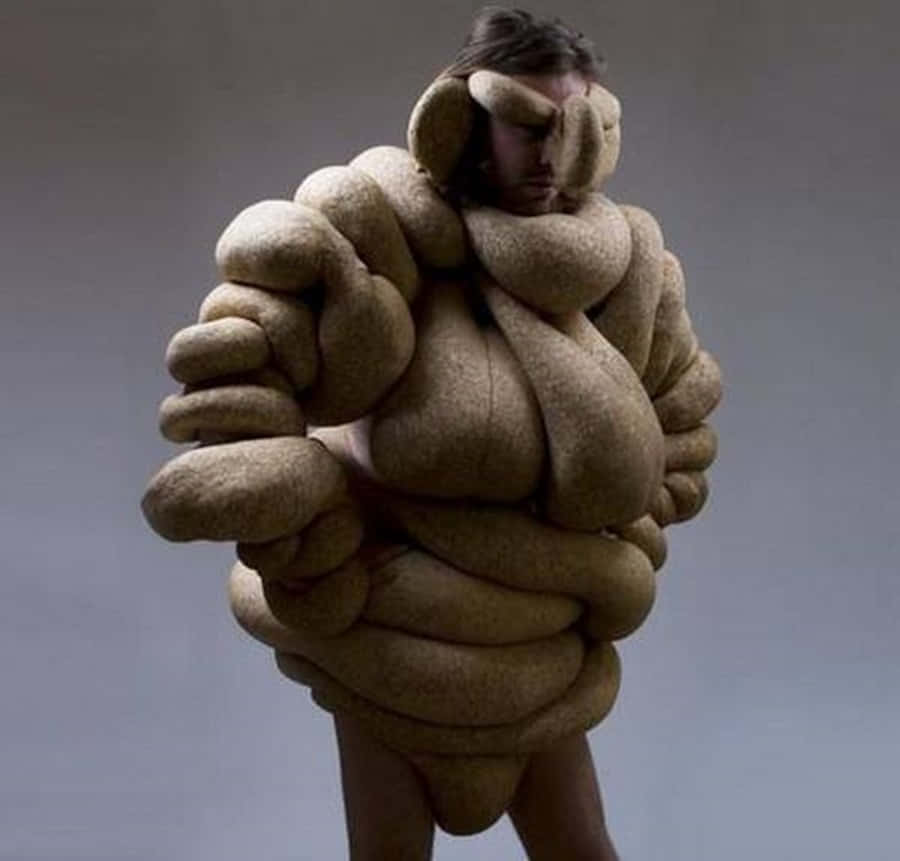 Freaky 桌布 壁紙 — 一個人被質感米色雕塑服裝包裹，類似扭曲的繩索或有機形態，背景為純灰色。