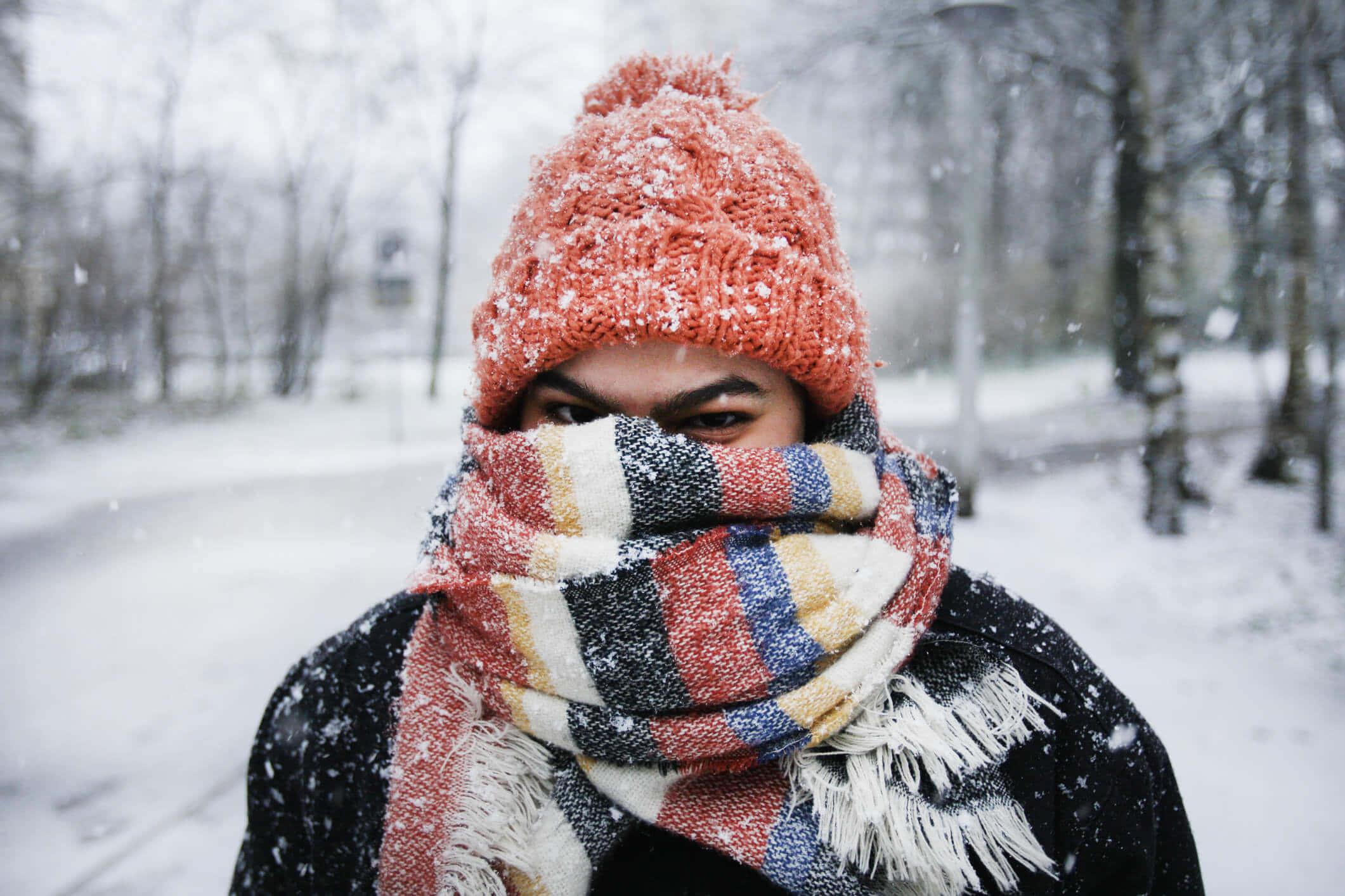 寒冷 桌布 壁紙 — 一個人裹著色彩繽紛的圍巾和沾滿雪的橙色針織帽，在冬季公園中透過飄落的雪花凝視著。