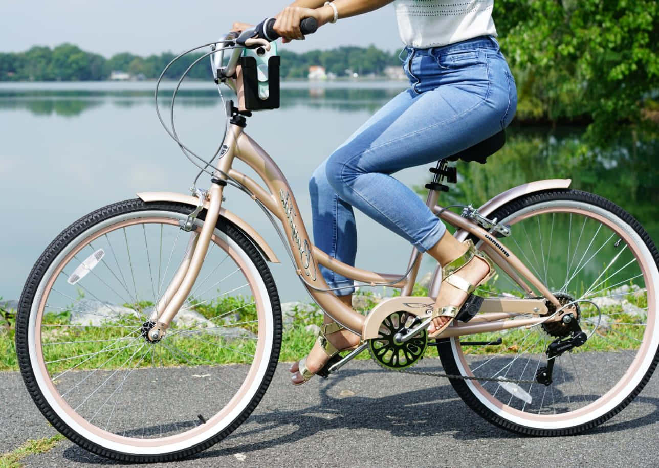 Beach Cruiser 桌布 壁紙 — 一個人騎著玫瑰金色單車沿著湖邊小徑行駛，背景是茂密的綠樹和寧靜的水面。