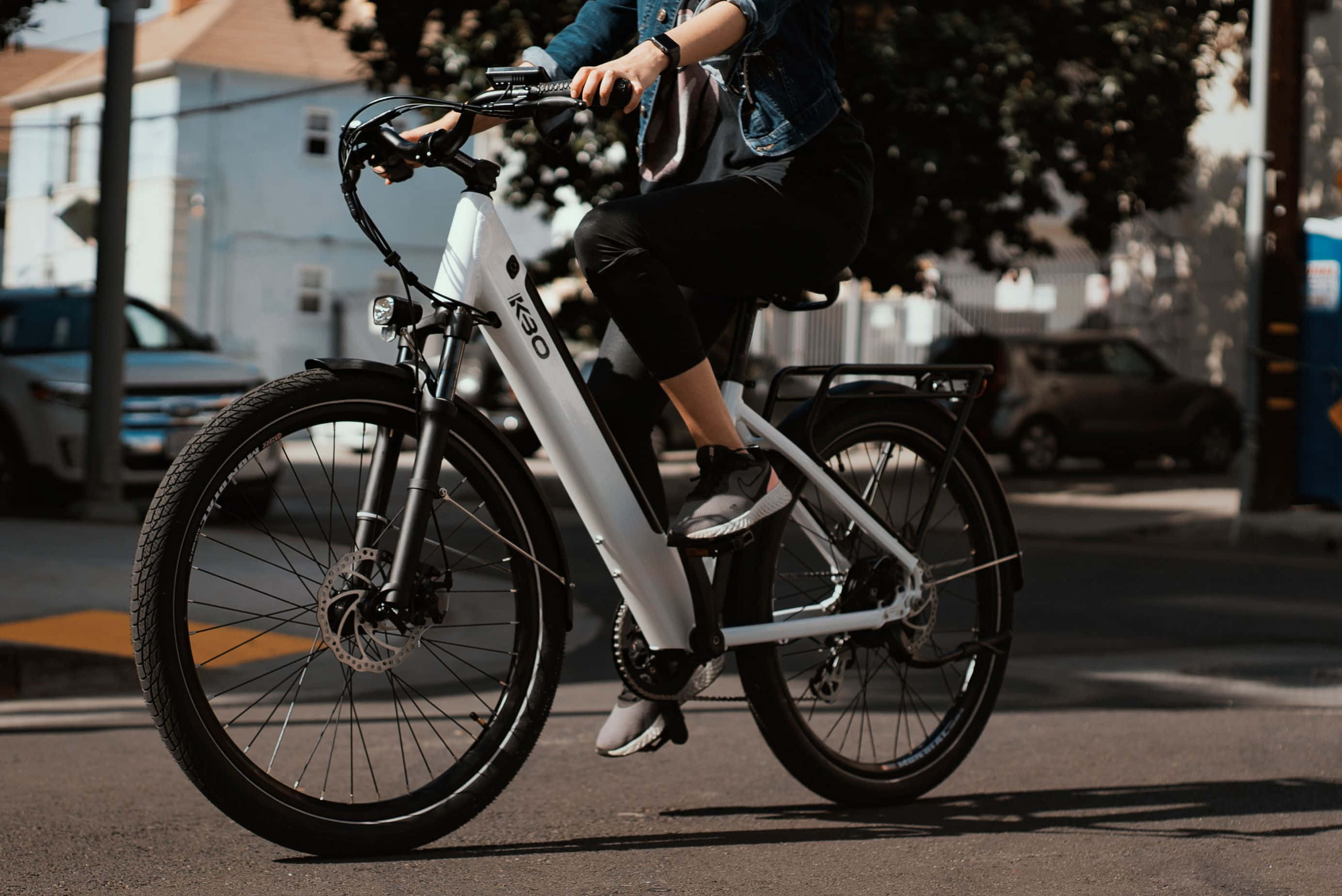Electric Bike 桌布 壁紙 — 一個人騎著白色電動單車，沿著陽光明媚的城市街道行駛，背景有停泊的汽車和樹木。